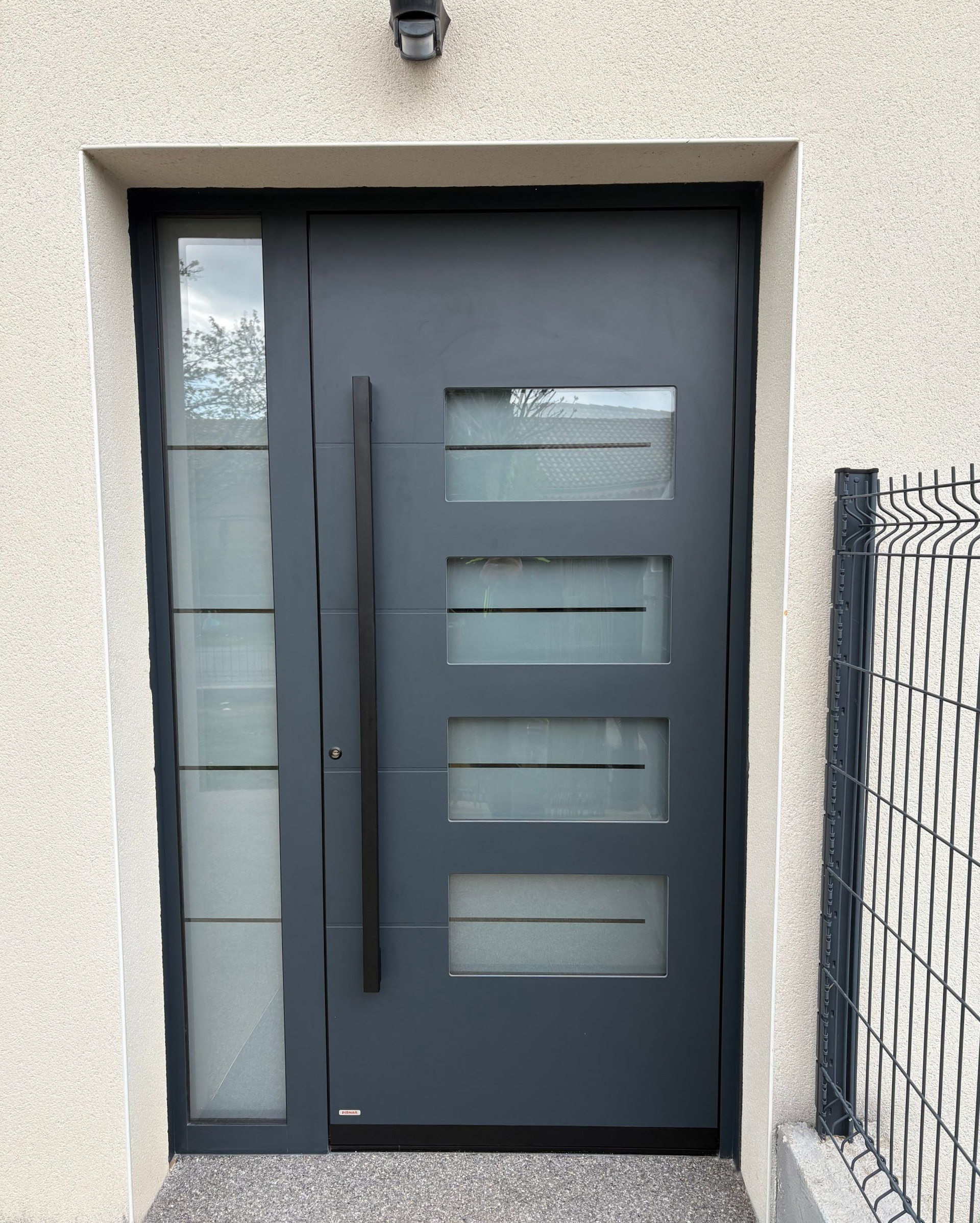 Porte d'entrée grise avec vitrae et une lôture grillagée sur le côté.