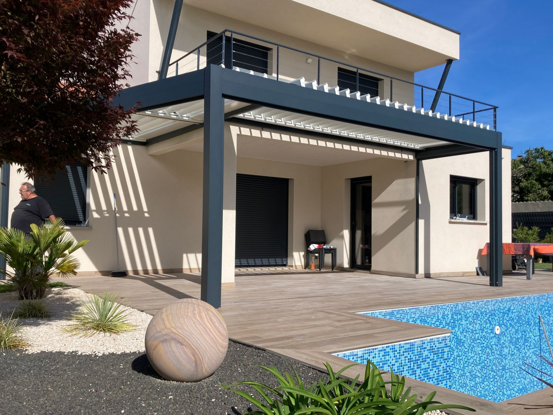 Une maison moderne avec une pergola à cadre sombre surplombant une terrasse au bord de la piscine, ornée d'une sphère en pierre et agrémentée d'un aménagement paysager.