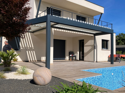 Une maison moderne avec une pergola en métal foncé, une terrasse en bois et une piscine dans un jardin ensoleillé.