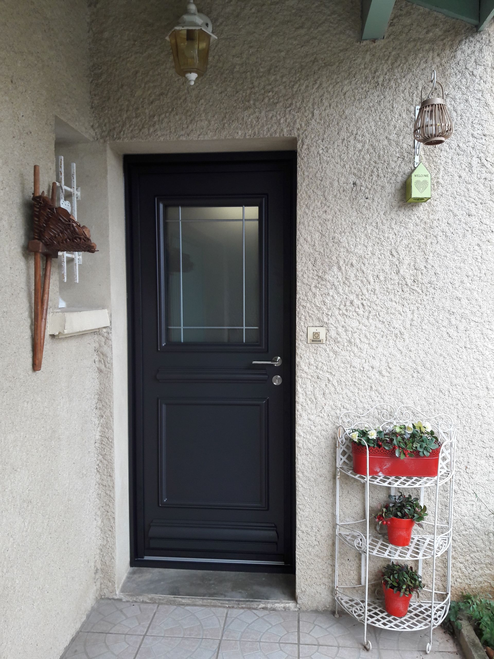 Une porte d'entrée noire avec un panneau vitré dépoli, encastrée dans un mur beige texturé, à côté d'un support à plantes à trois niveaux.