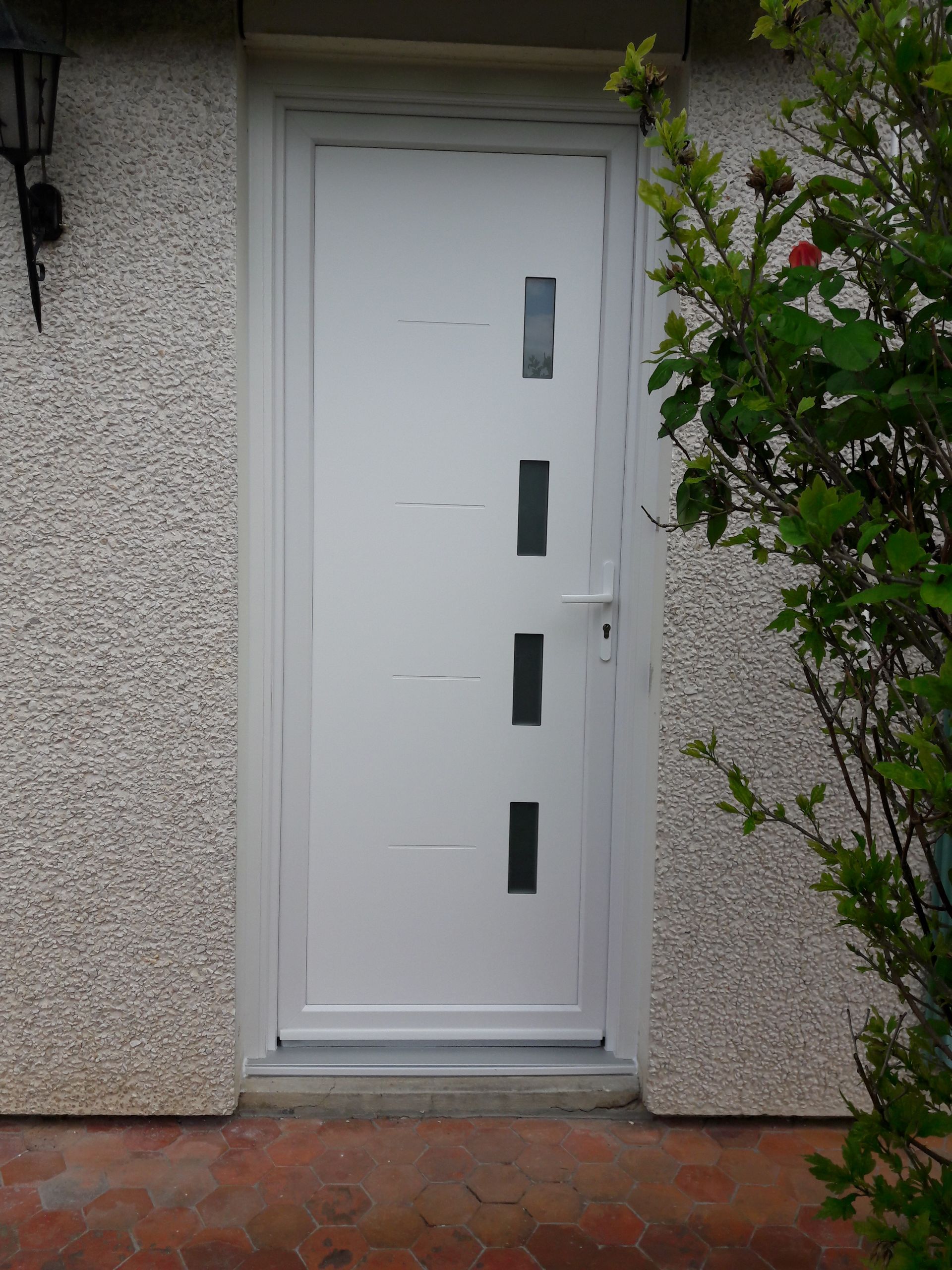 Une porte extérieure blanche avec quatre petits carreaux de verre rectangulaires verticaux, encastrée dans un mur en crépi clair.