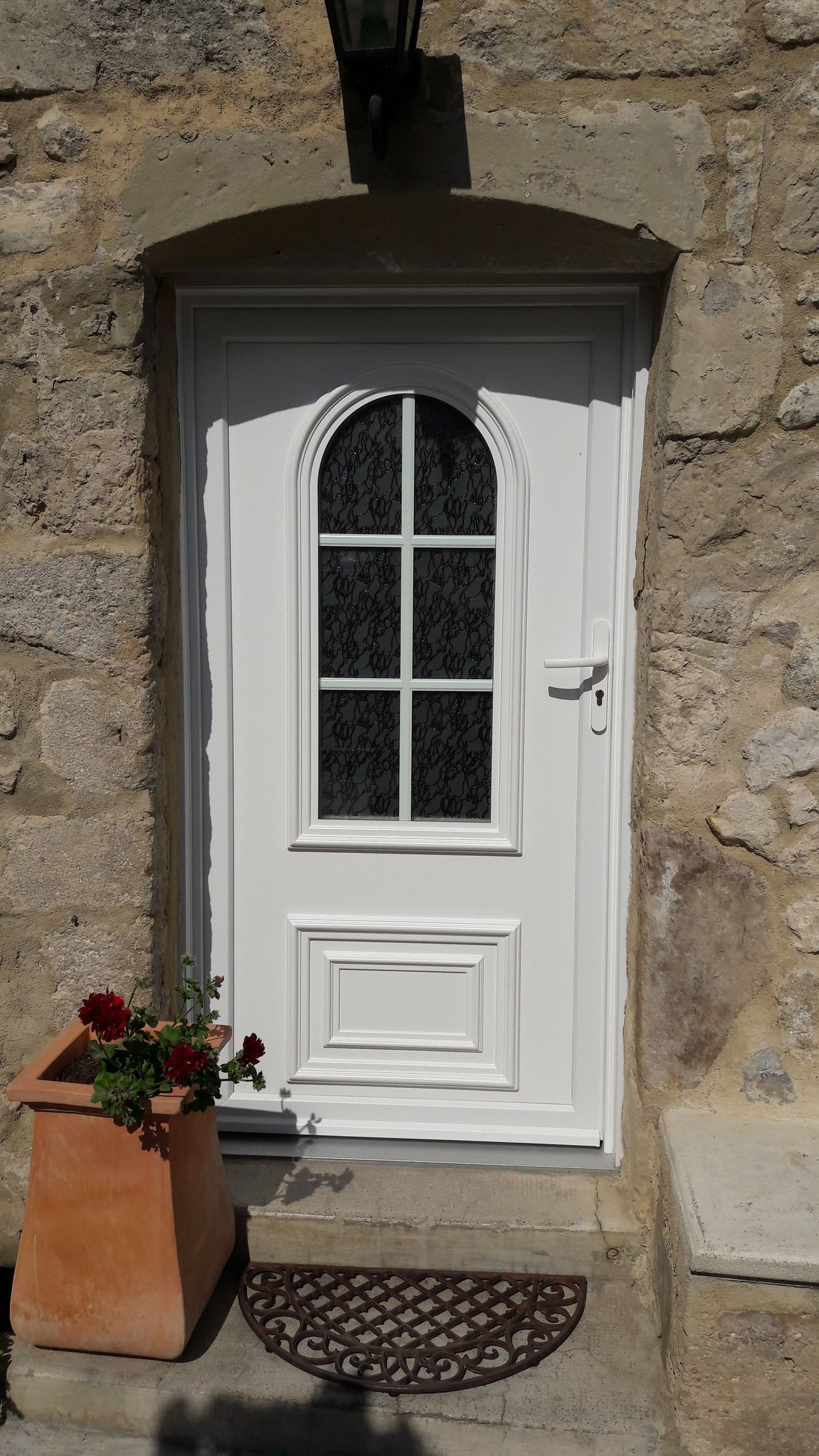 Une porte d'entrée blanche avec une vitre, encastrée dans un mur de pierre, avec une plante rouge en pot et un paillasson sur le seuil.