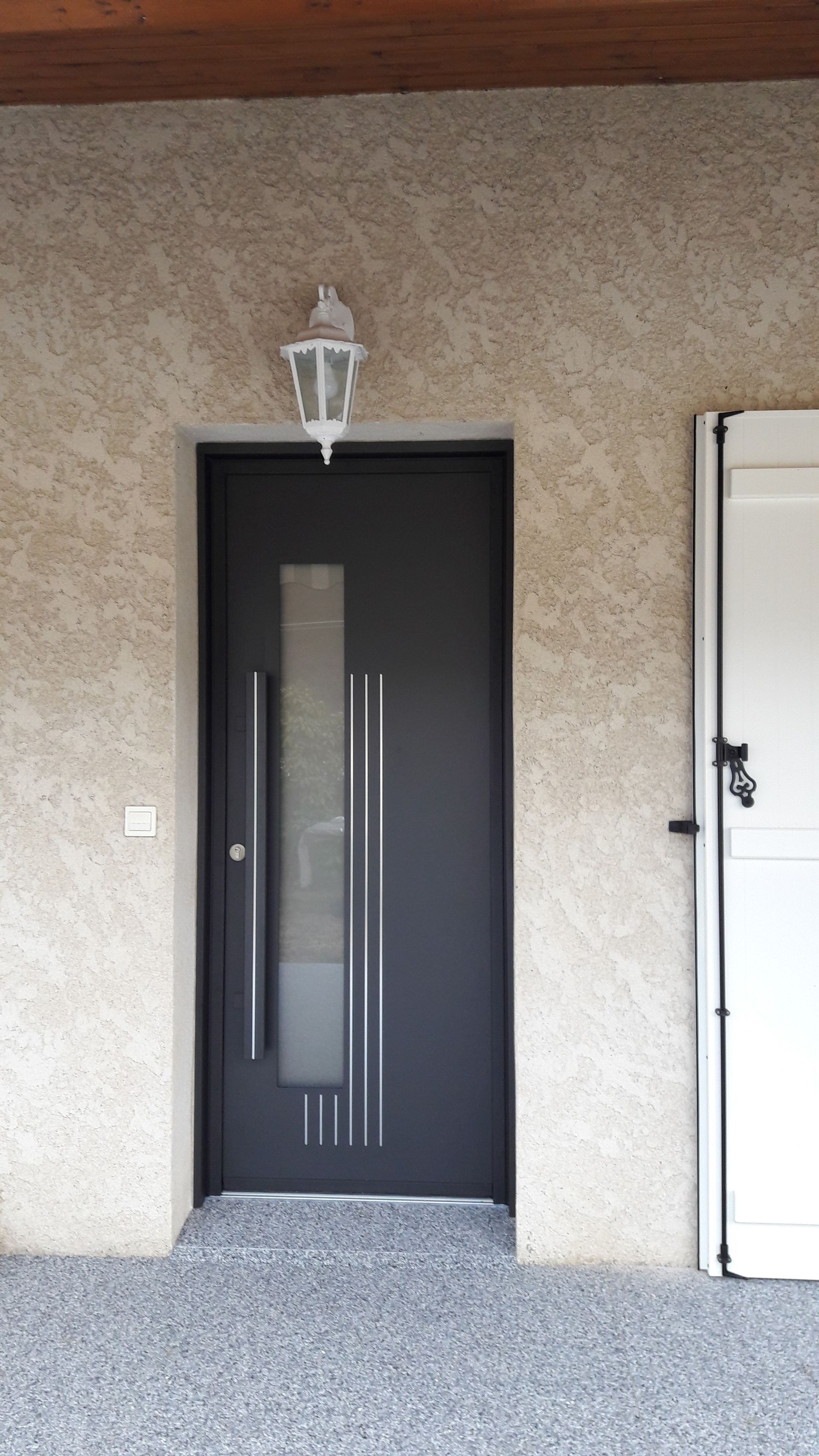 Une porte d'entrée gris foncé avec un panneau de verre vertical et une poignée en métal moderne, encastrée dans un mur extérieur en stuc clair.