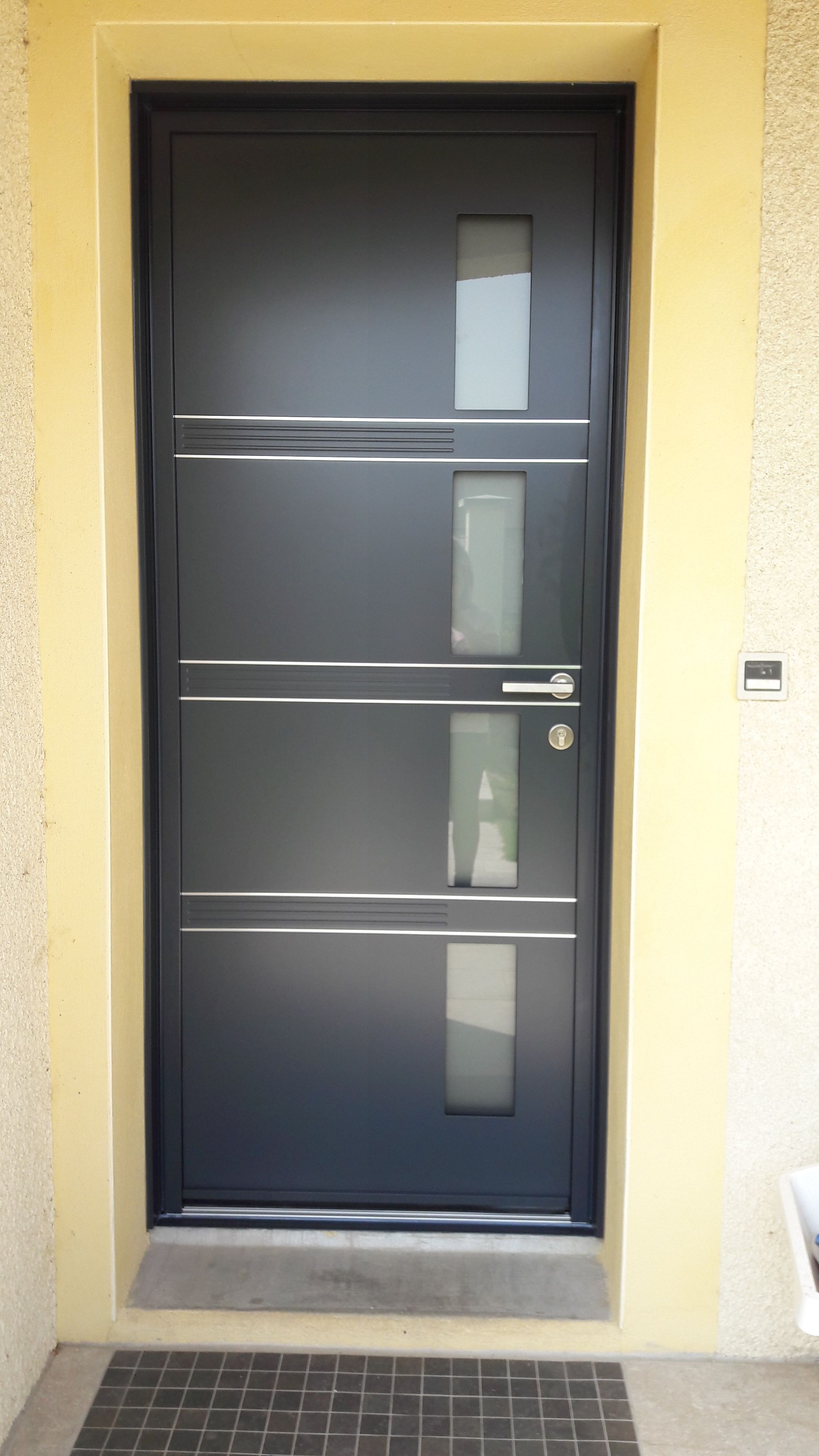 Une porte d'entrée rectangulaire gris foncé, ornée de quatre fines fenêtres verticales en verre dépoli, s'insère dans un hall d'entrée jaune clair.