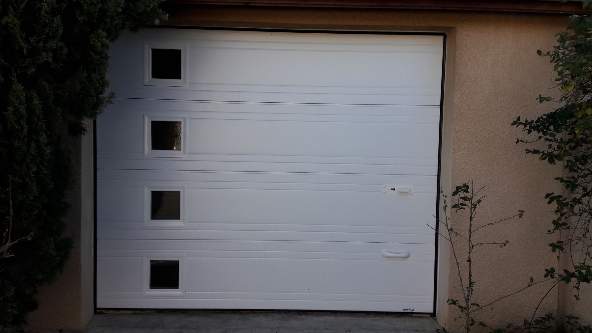 Une porte de garage blanche comportant une colonne verticale de quatre fenêtres carrées sur le côté gauche, intégrée à un mur résidentiel.