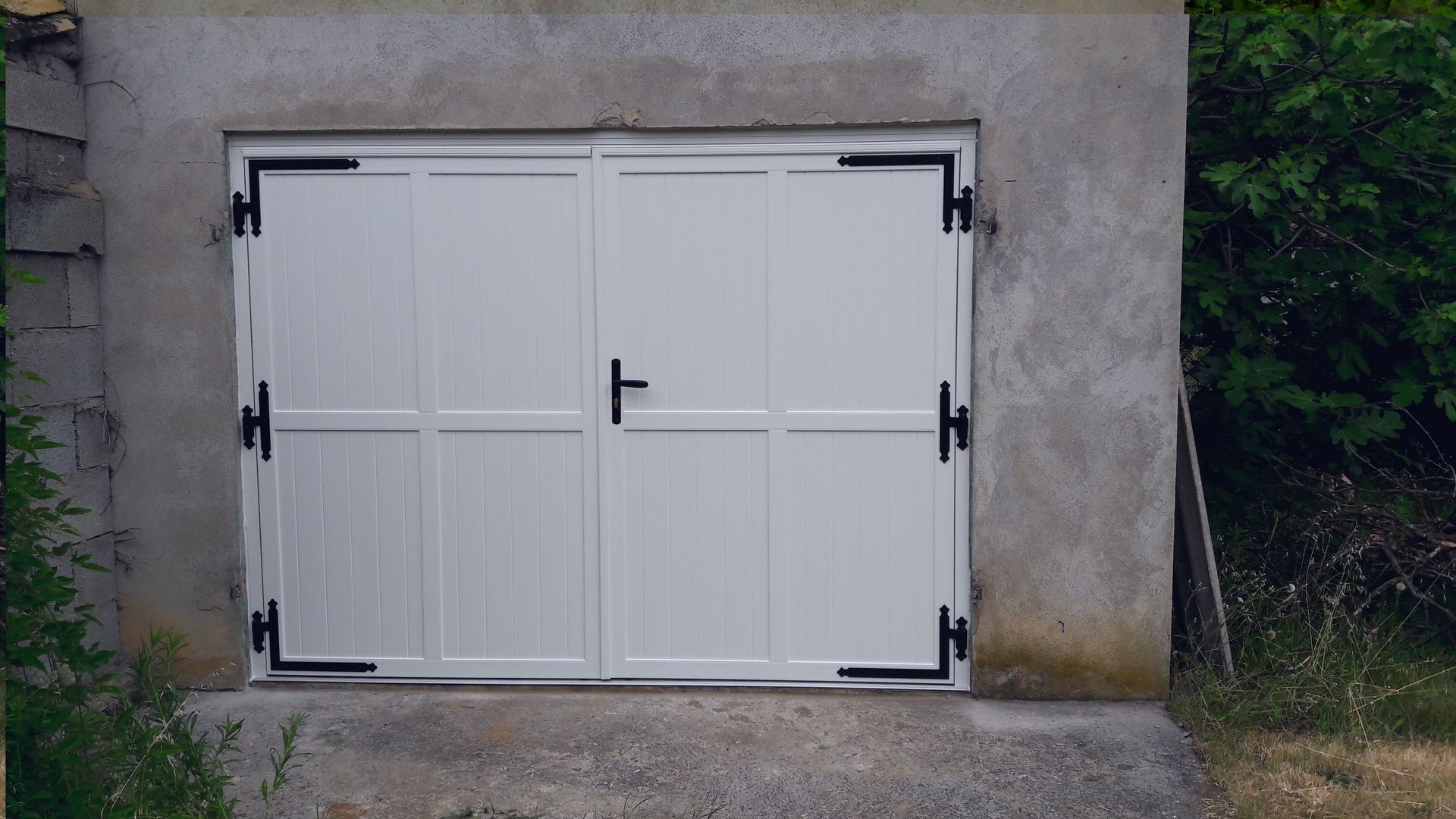 Porte de garage double blanche avec charnières noires et poignée centrale, encastrées dans un mur en béton brut.