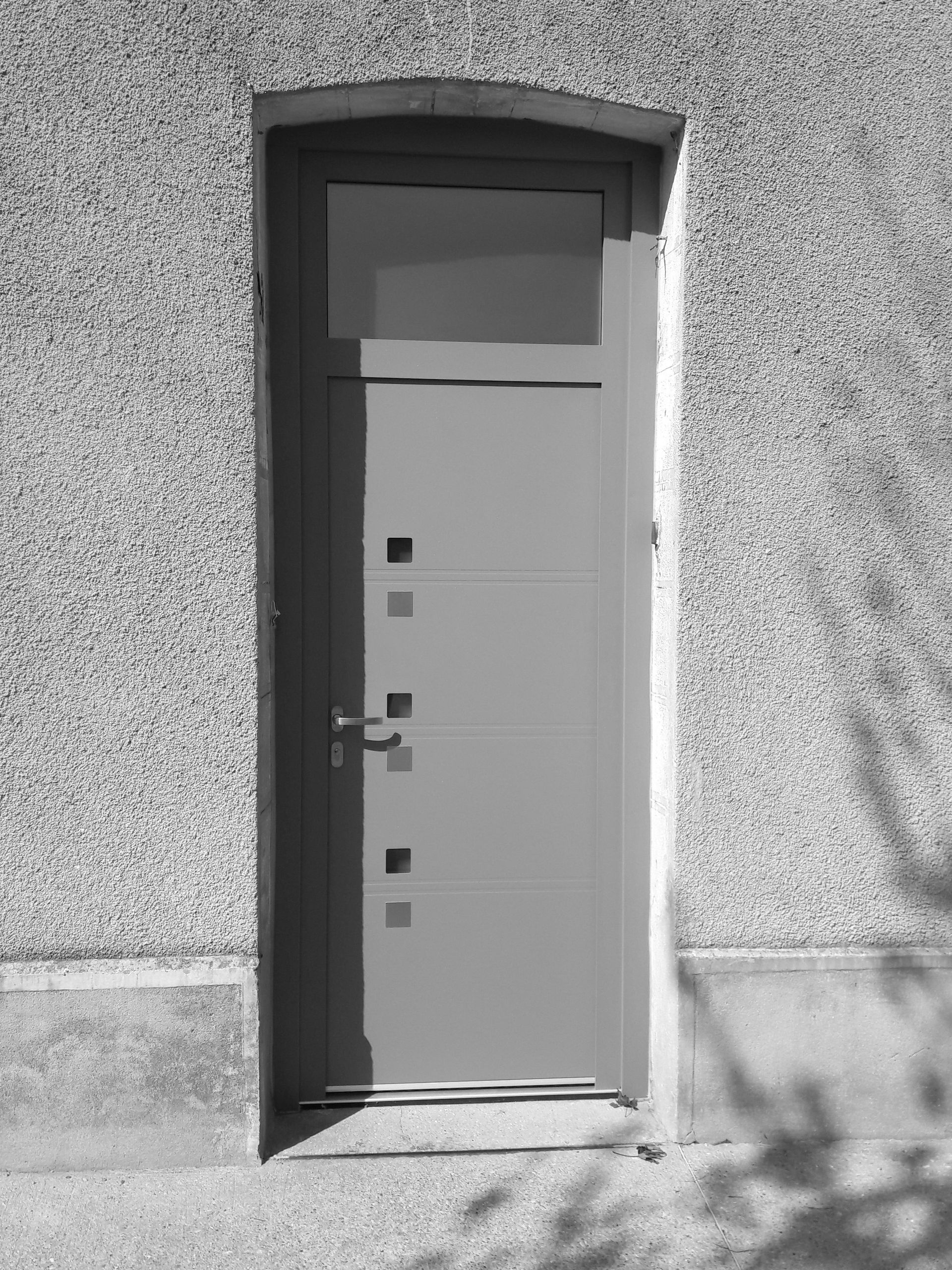 Une porte rectangulaire grise avec une vitre en partie supérieure et quatre petits éléments décoratifs carrés, encastrée dans un mur texturé.
