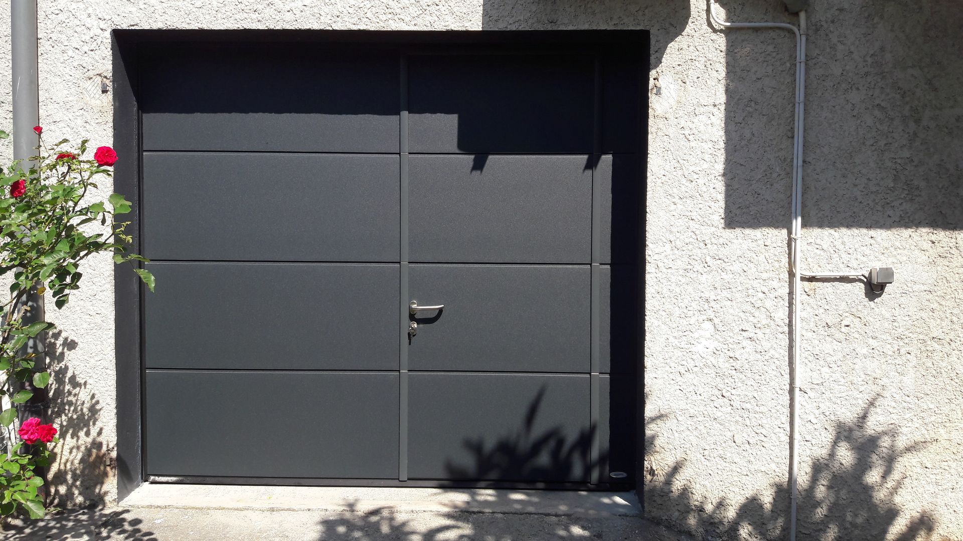 Une porte de garage moderne gris foncé à panneaux horizontaux, encastrée dans un mur de pierre texturée à côté d'un rosier grimpant.