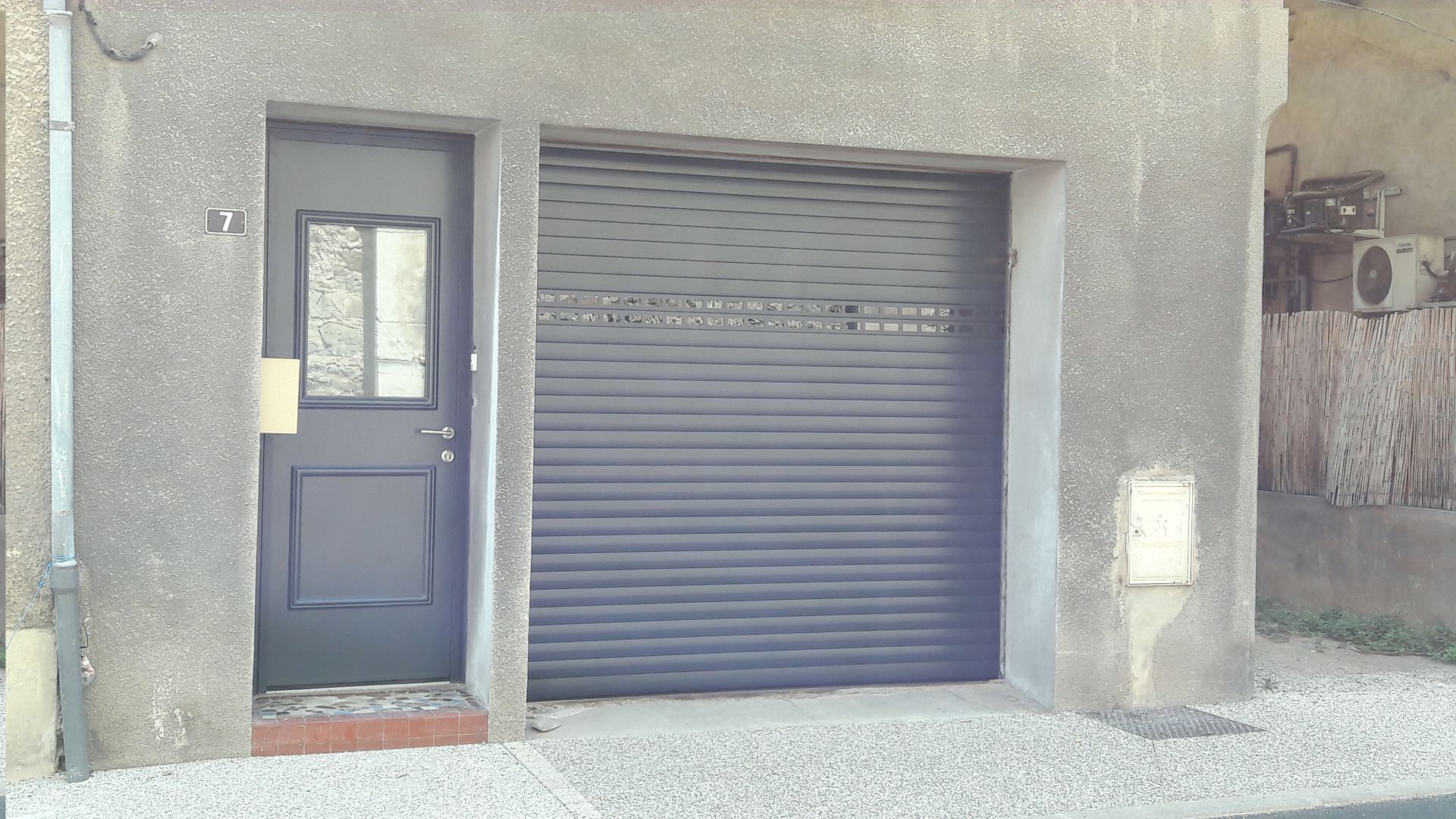 La façade grise d'un bâtiment présente une porte sombre avec une vitre, jouxtant une porte de garage coulissante horizontale assortie.