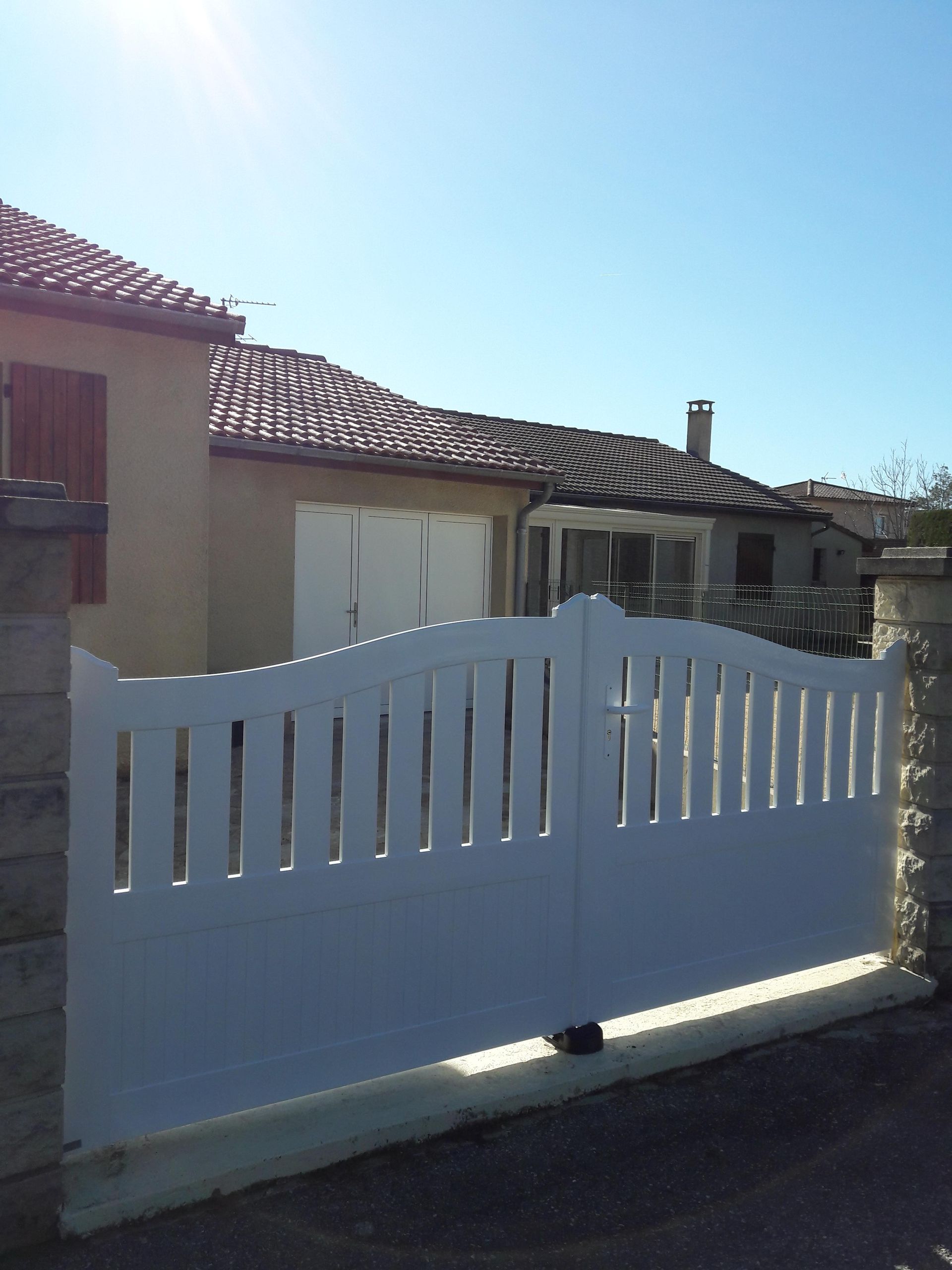 Un portail métallique blanc en forme d'arche, orné de lattes verticales, se dresse devant une maison d'habitation au toit de tuiles, sous un ciel ensoleillé.
