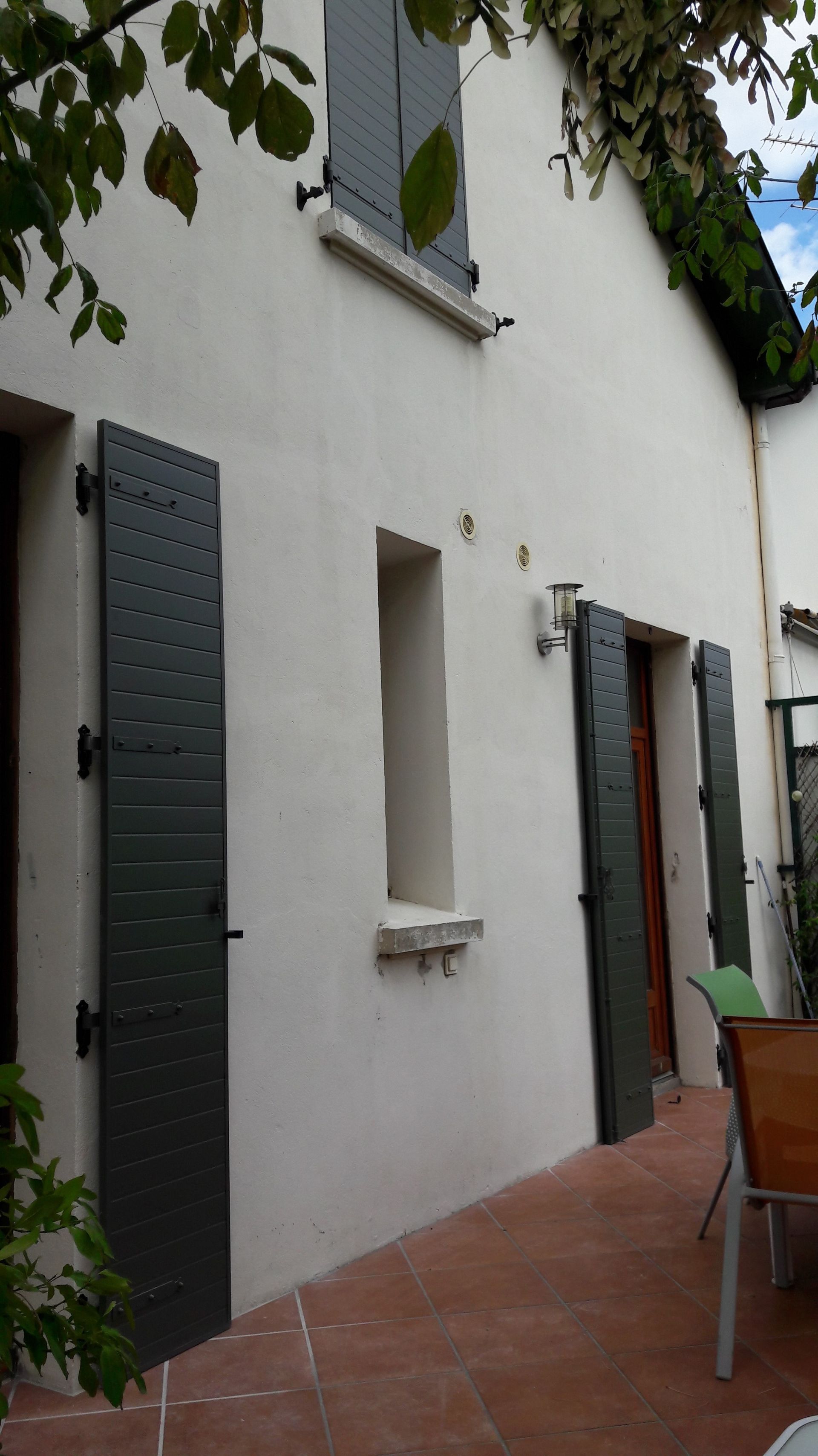 Une maison à l'extérieur en stuc blanc avec des volets vert foncé, donnant sur un patio ensoleillé carrelé de terre cuite.
