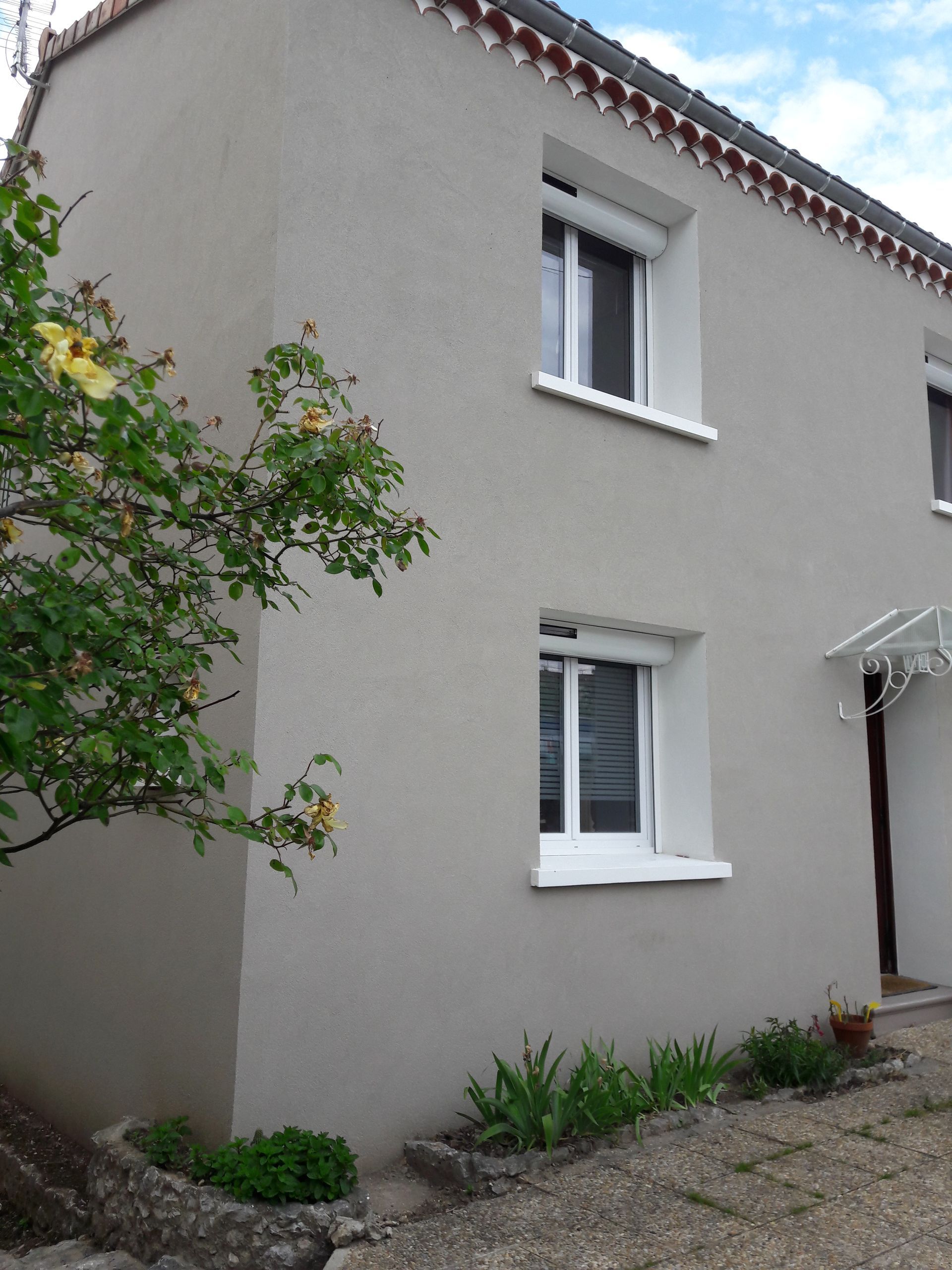 Maison à deux étages avec des murs en stuc gris clair, une bordure de toit en tuiles brunes et un petit rosier dans la cour avant.