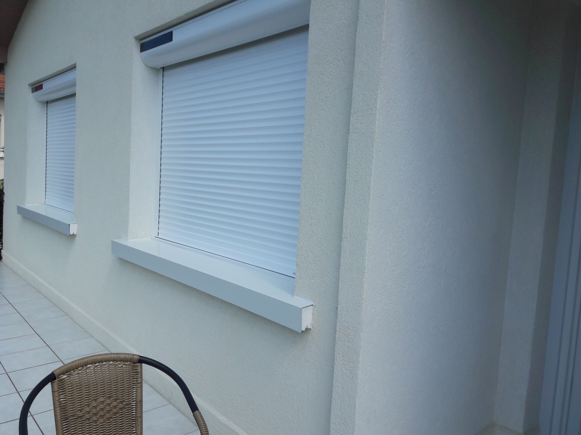 Mur blanc d'une maison avec deux fenêtres munies de volets roulants solaires clairs fermés, au-dessus d'une terrasse carrelée.