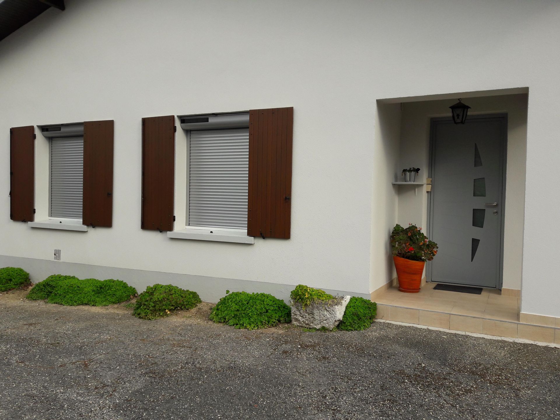 L'extérieur d'une maison blanche présente deux fenêtres avec des volets et des stores enrouleurs marron, à côté d'une porte grise avec une plante en pot.