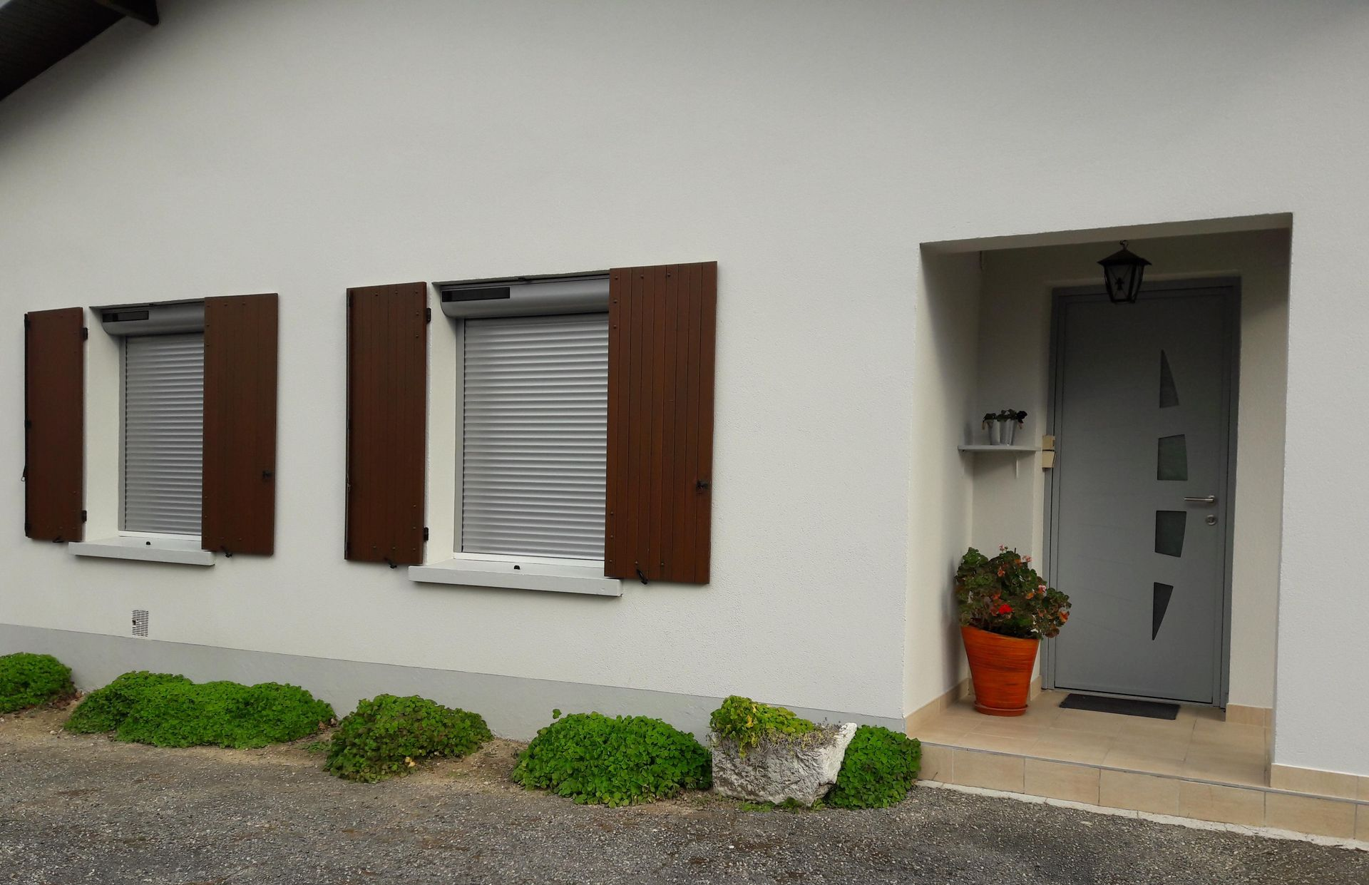 Vue extérieure d'une maison blanche avec deux fenêtres à volets marron, une porte d'entrée grise et des arbustes verts au niveau du sol.