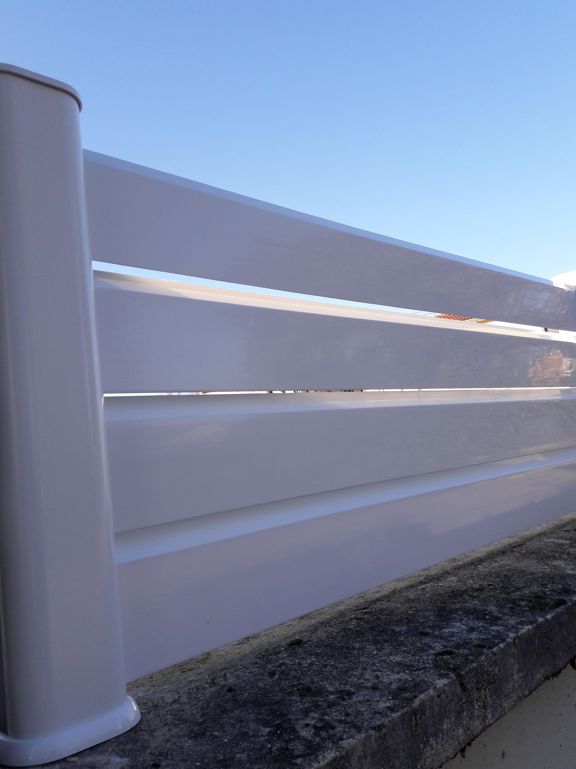 Panneaux de clôture horizontaux blancs, fixés par un poteau vertical, montés sur un mur de pierre se détachant sur un ciel bleu clair.
