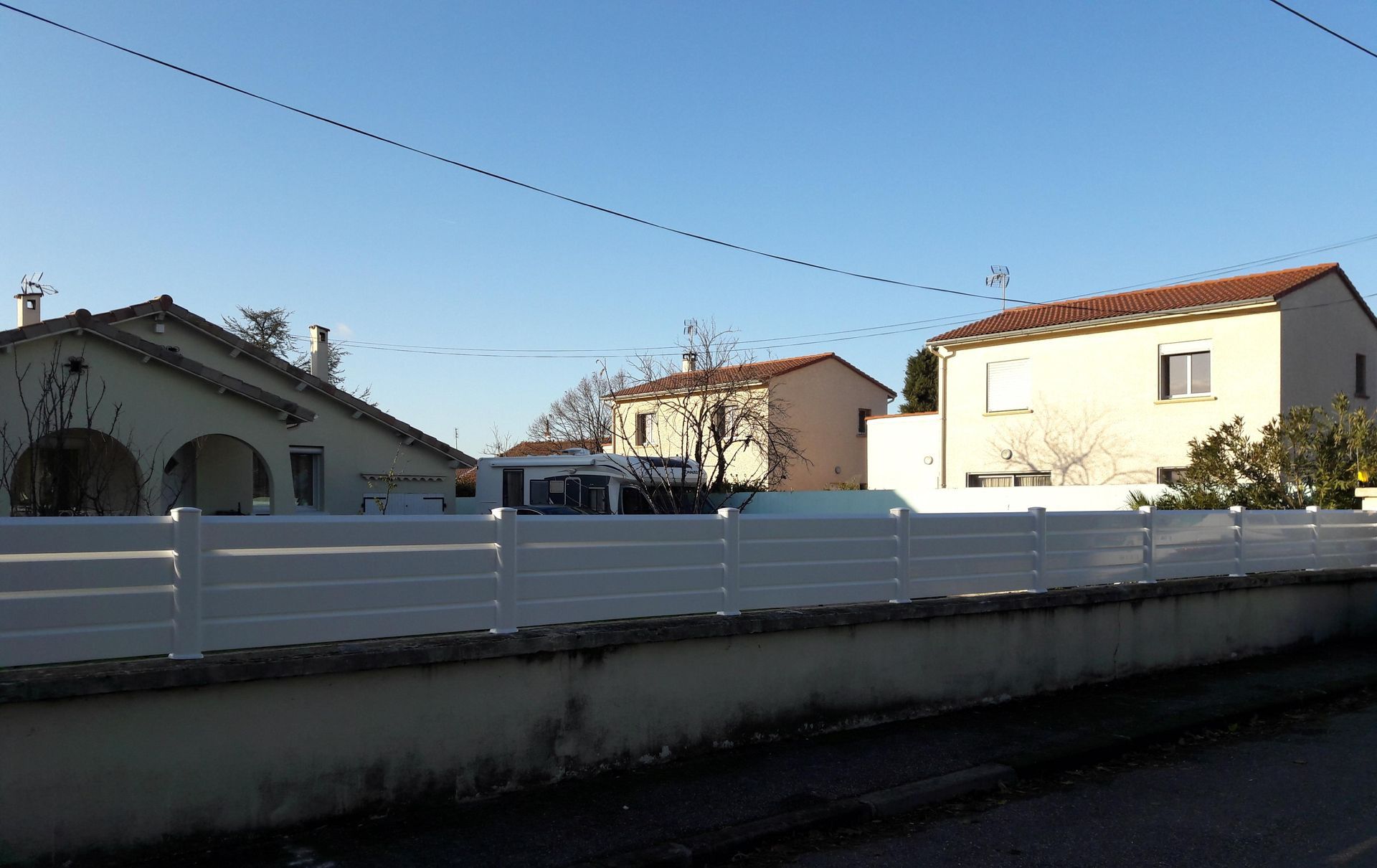 Une clôture blanche horizontale se dresse devant plusieurs maisons sous un ciel bleu limpide.