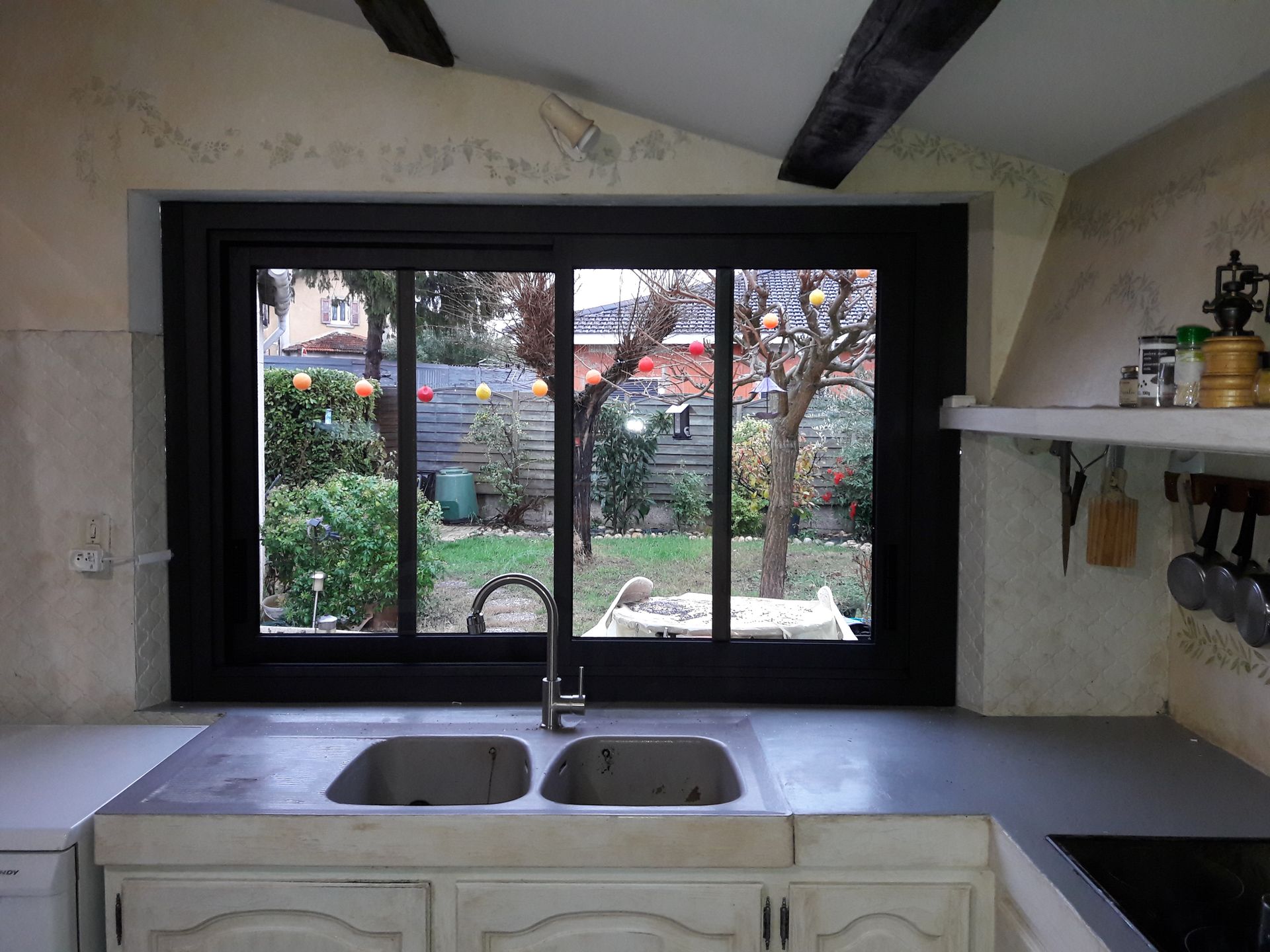 Un évier de cuisine à double bac est installé sous une fenêtre à cadre noir donnant sur un jardin arboré à l'arrière.