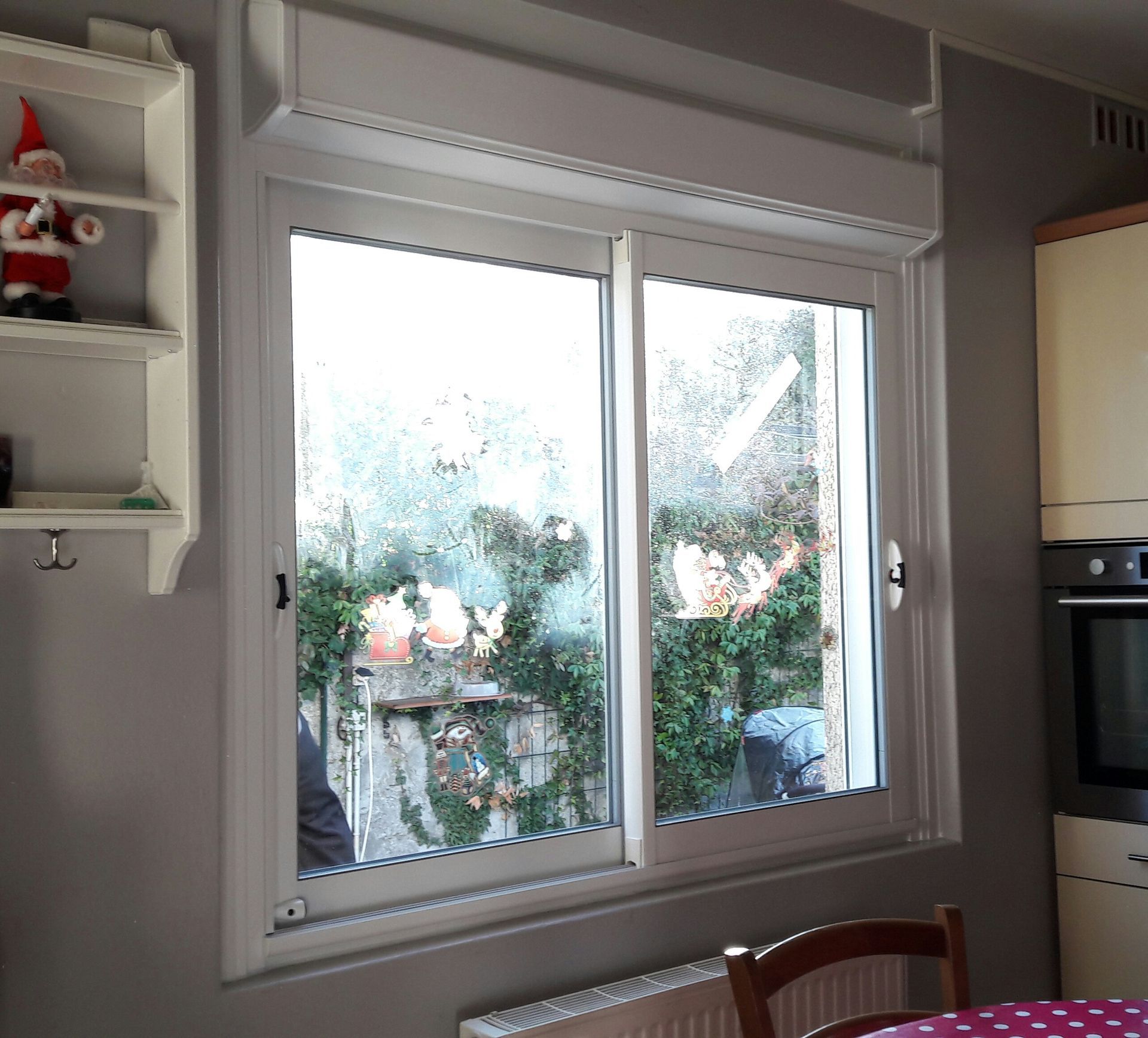 Une fenêtre avec un volet roulant blanc installé au-dessus, située dans une pièce avec une étagère et un appareil électroménager sur le côté.
