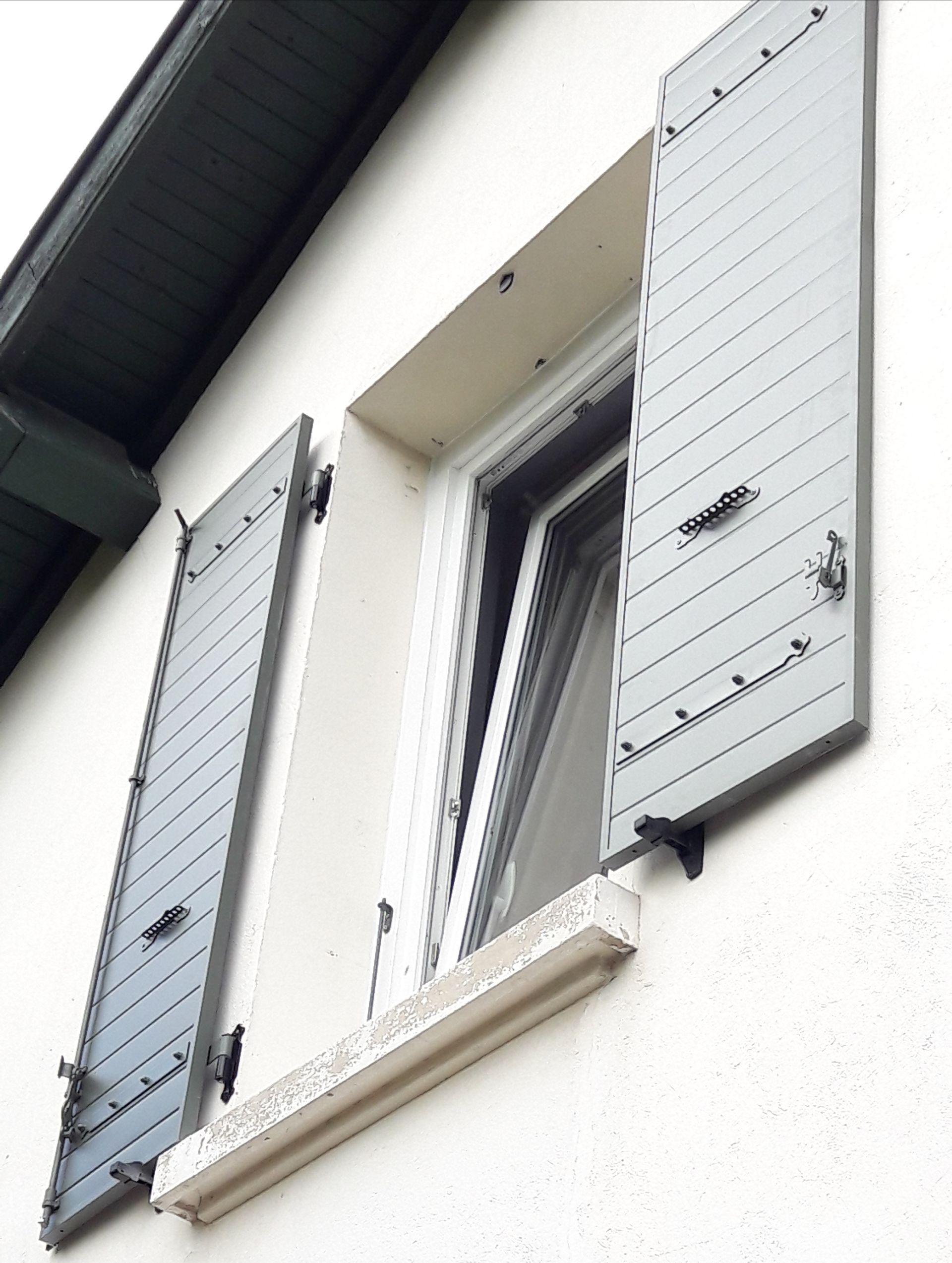 Une fenêtre à volets gris clair sur un mur extérieur blanc, vue d'un angle bas.