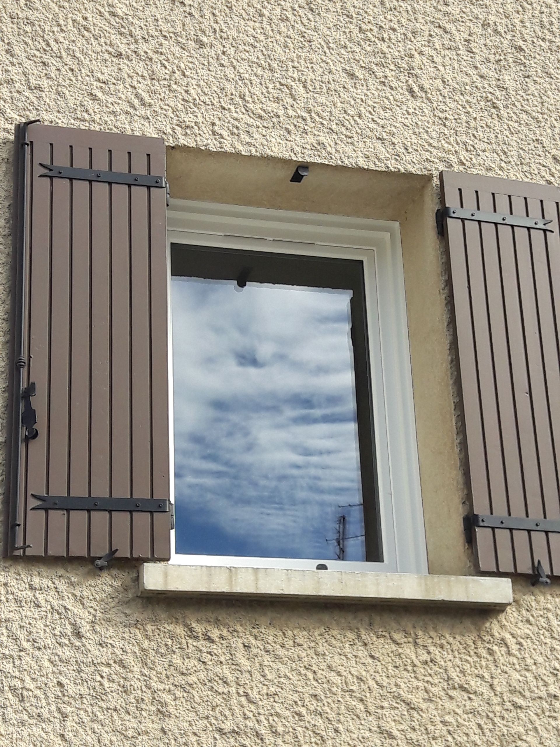Une fenêtre rectangulaire avec des volets en bois brun, installée sur un mur extérieur en stuc texturé de couleur claire.