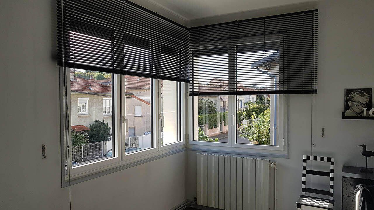 Fenêtres d'angle avec stores vénitiens noirs, un radiateur blanc en dessous et un petit cadre photo sur le mur de droite.