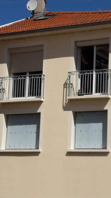 Façade d'immeuble beige avec deux niveaux de fenêtres, deux balcons avec garde-corps métalliques et un toit de tuiles rouges avec une antenne parabolique.