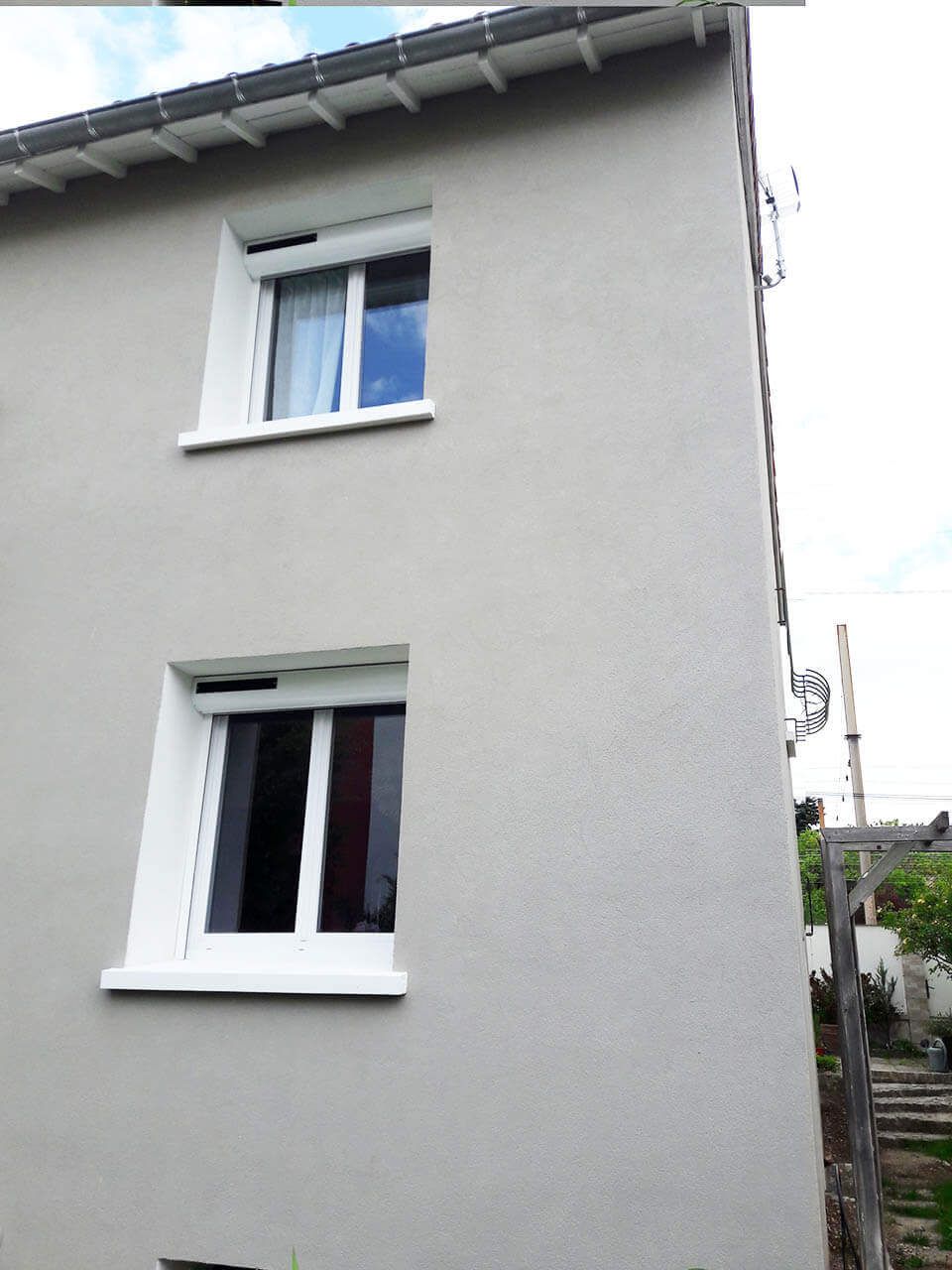 L'extérieur d'une maison à deux étages, de couleur gris clair, avec deux fenêtres à cadre blanc, surmontées de caissons de volets roulants visibles.