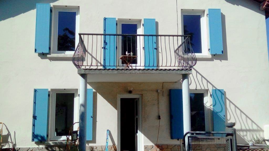 Une maison à deux étages avec des murs blancs, des volets bleus et un balcon au-dessus de l'entrée principale.