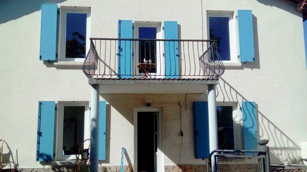 Une maison à deux étages aux murs blancs, aux volets bleus et au balcon central soutenu par deux piliers au-dessus de l'entrée.
