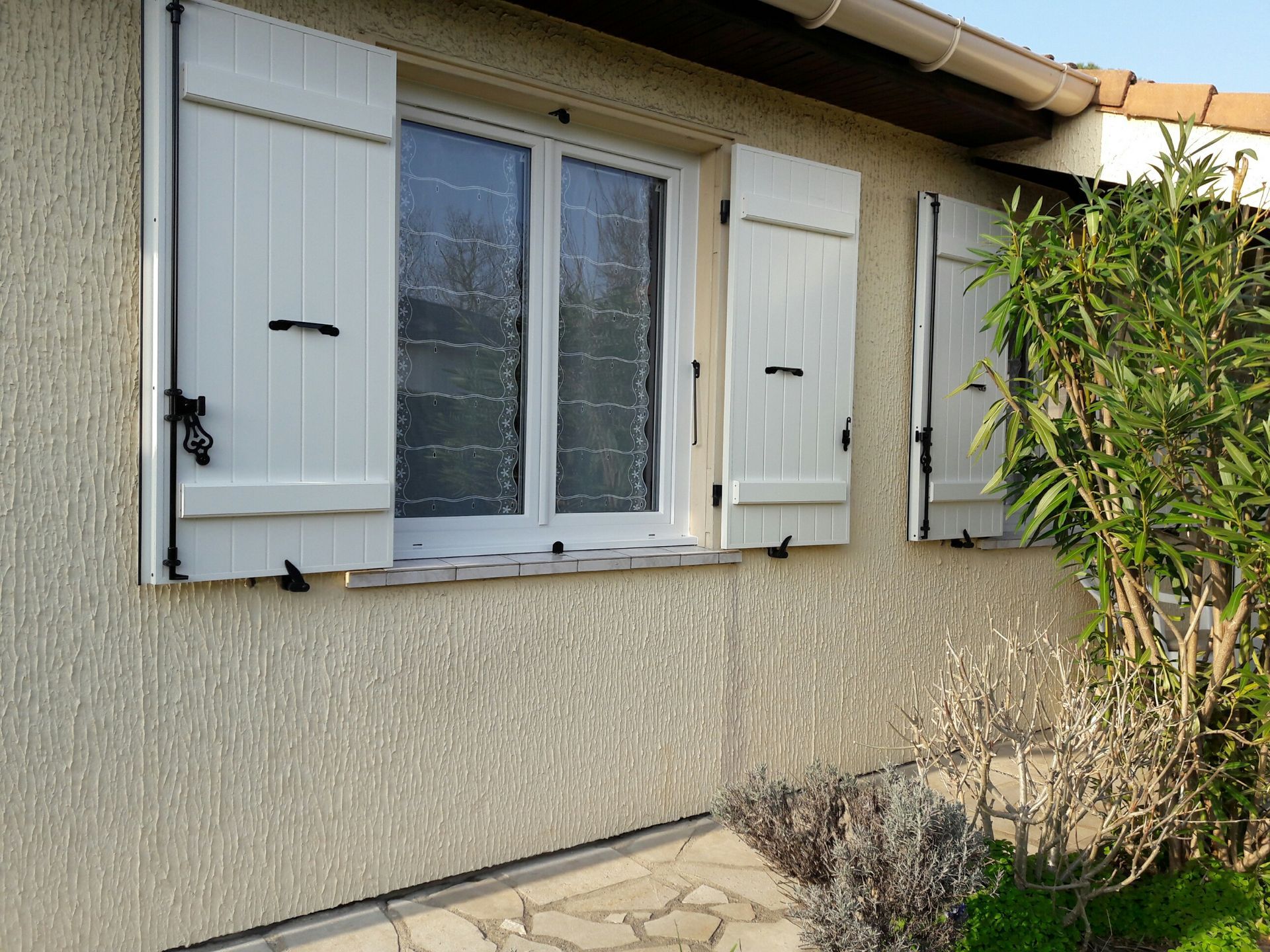 Vue extérieure d'un mur de maison beige texturé, avec une fenêtre à volets blancs et un petit buisson sur la droite.
