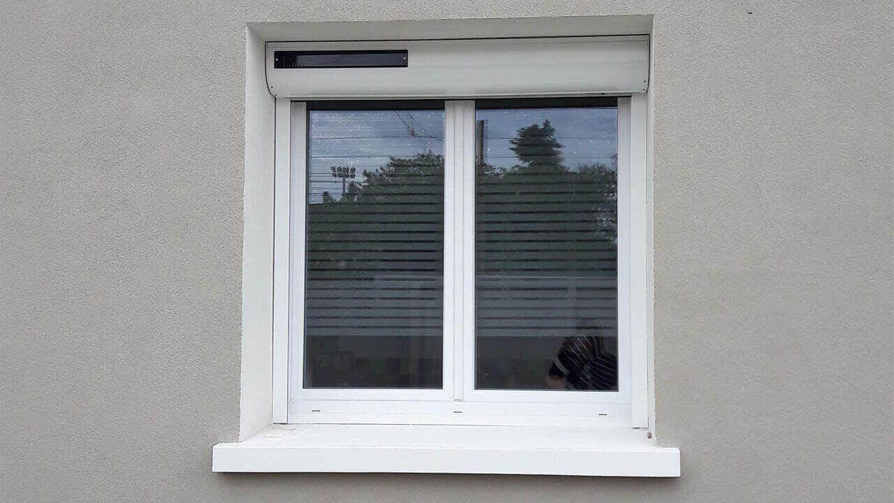 Une fenêtre à cadre blanc avec un coffre de volet roulant fermé sur un mur en stuc gris.