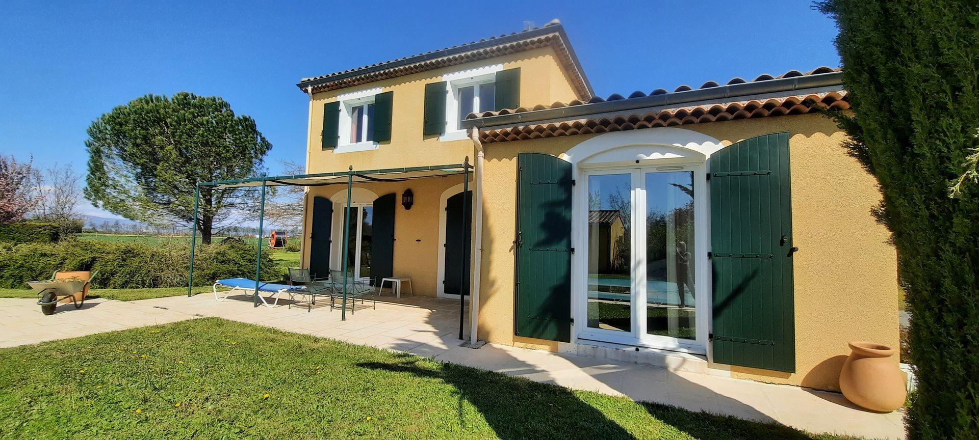 Une maison à deux étages en stuc jaune avec des volets verts et une terrasse à l'avant, située dans un jardin ensoleillé avec un petit chien.