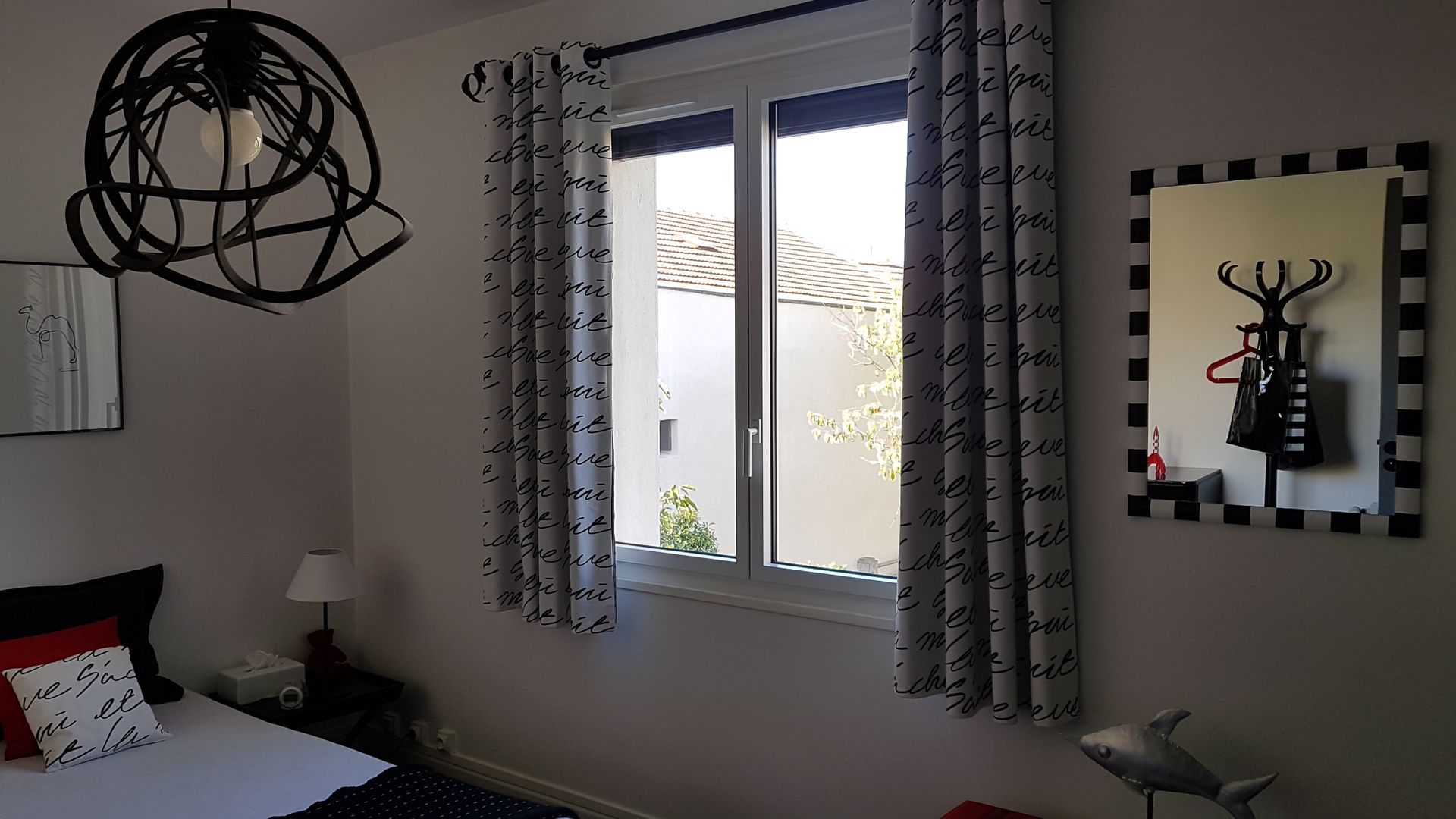 Une chambre avec un luminaire unique en fil de fer noir au design abstrait, des rideaux à motifs, une lampe de chevet et un miroir mural.