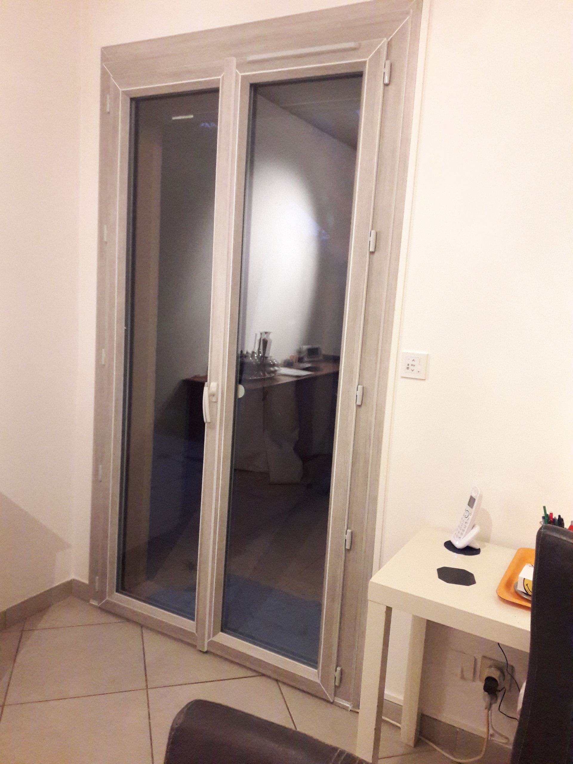 Une porte-fenêtre à double vitrage avec un cadre en bois clair, située dans une pièce au sol carrelé et avec une petite table.