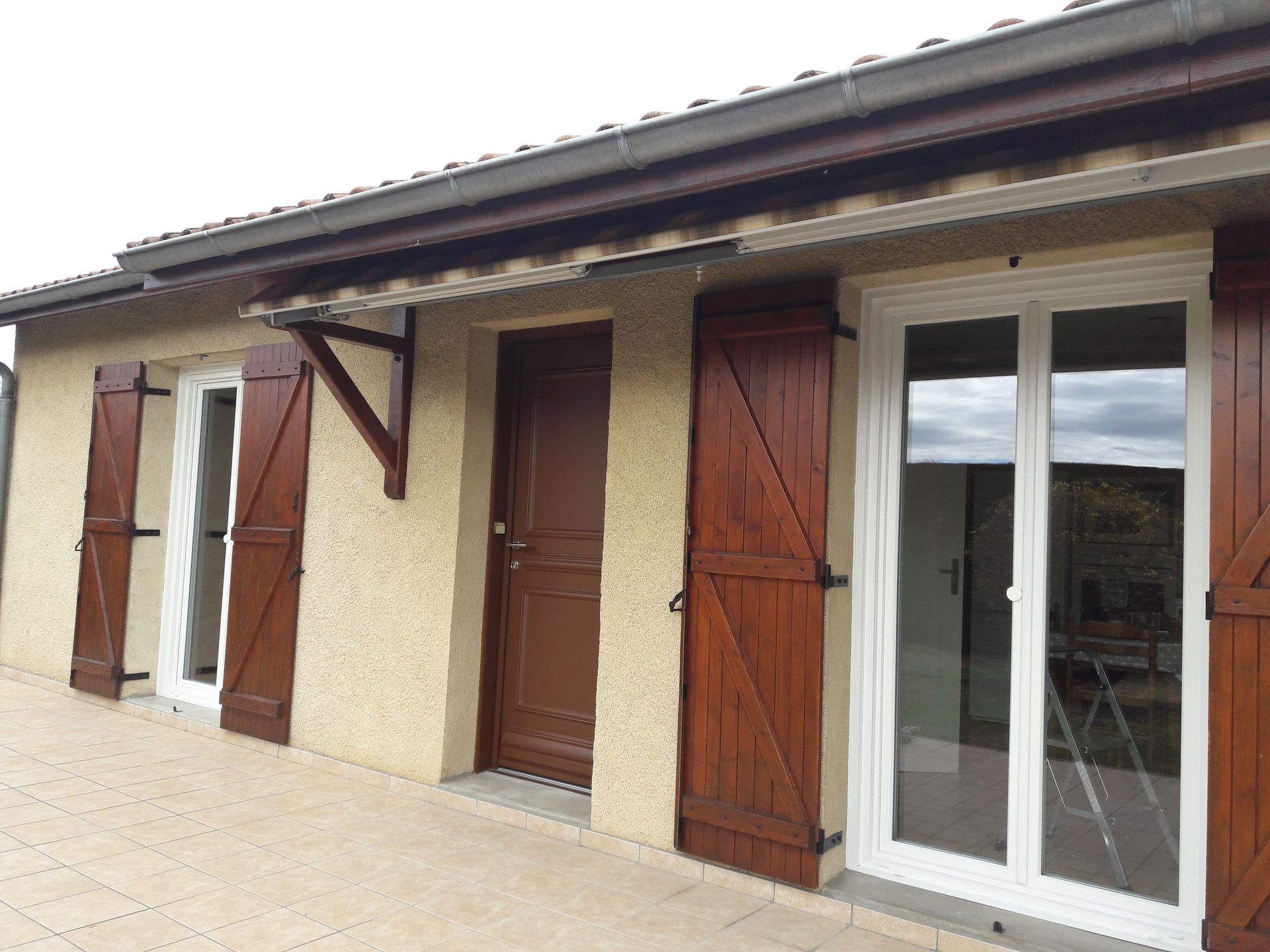 Extérieur d'une maison beige avec une terrasse pavée, des volets en bois, une porte en bois massif et une porte coulissante en verre blanc.