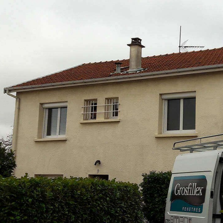 Une maison beige à deux étages avec un toit de tuiles rouges, comportant deux grandes fenêtres et deux petites fenêtres à barreaux au centre.