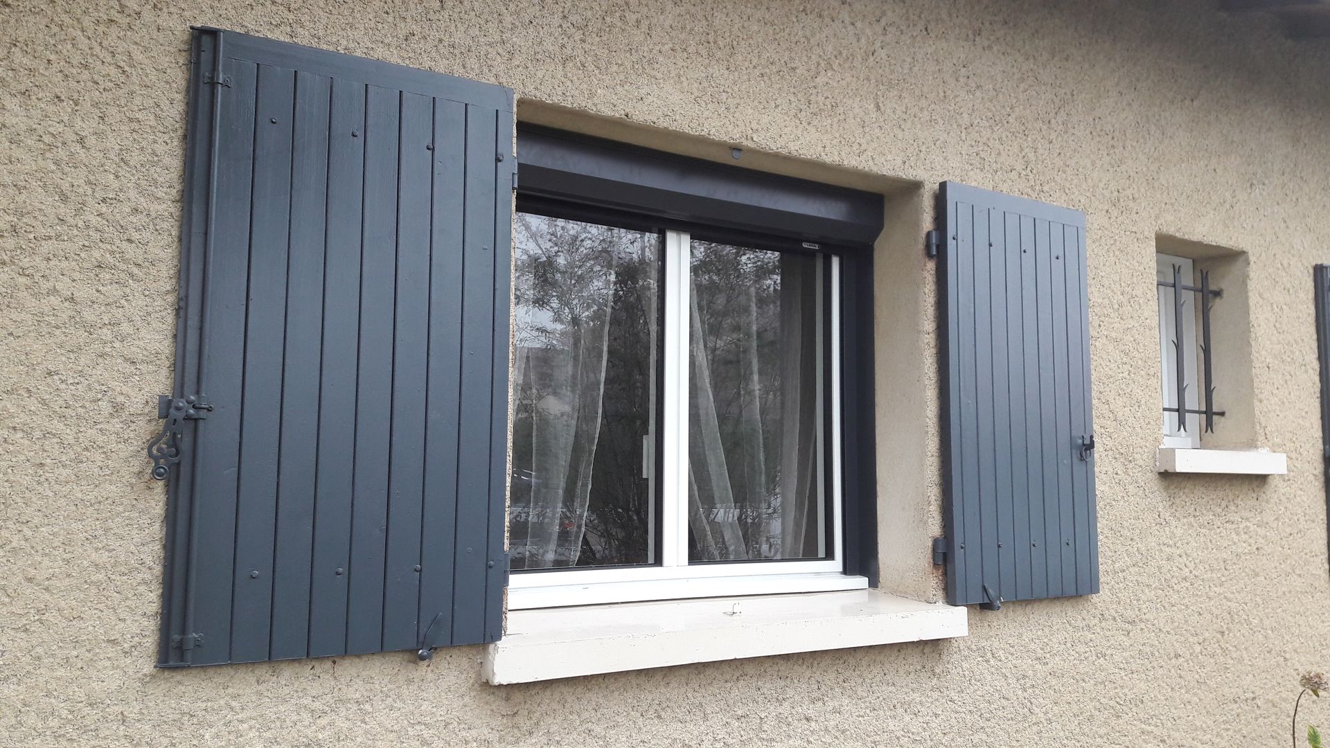 Une fenêtre à cadre sombre avec des volets verticaux ouverts, gris ardoise, encastrée dans un mur extérieur beige en crépi à la gravillons.