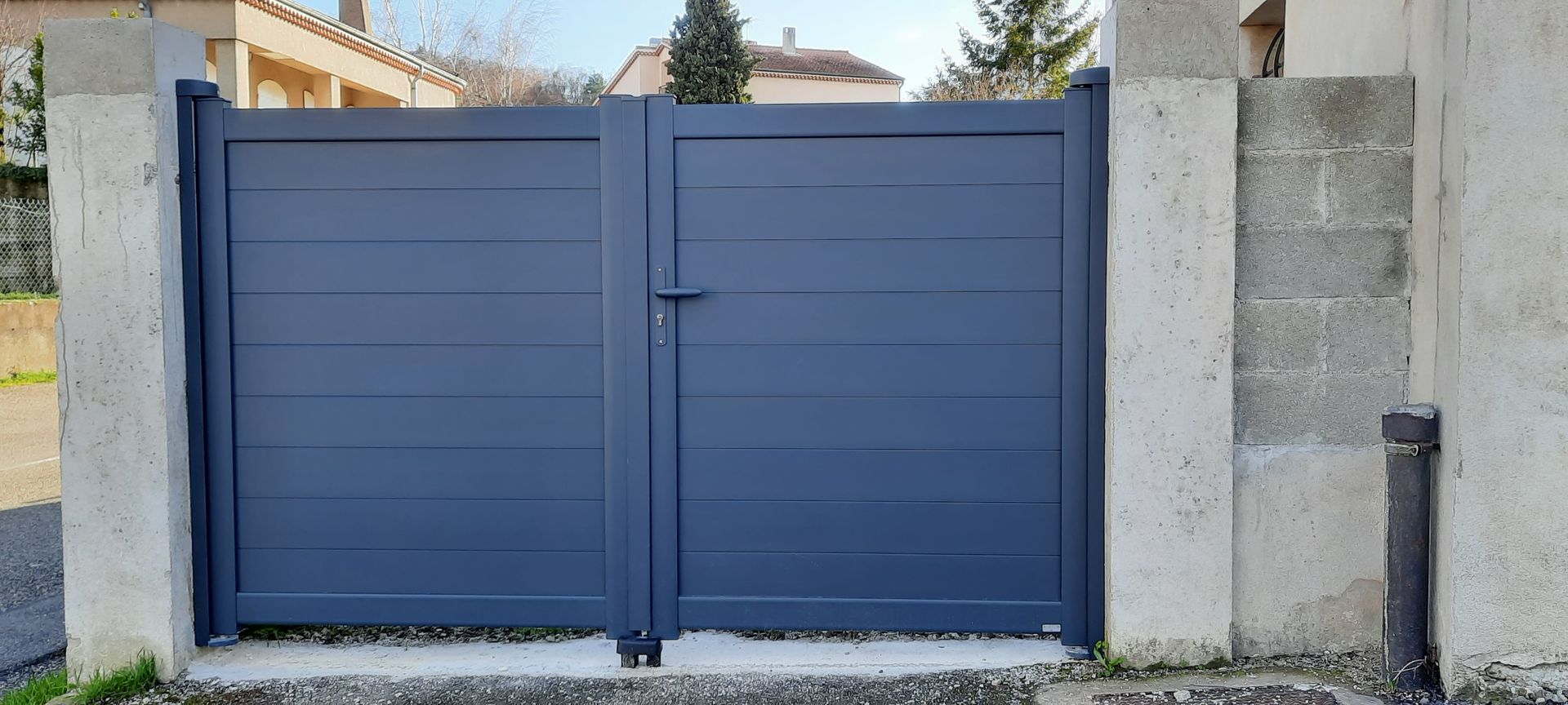 Un portail double moderne, bleu foncé, à panneaux pleins, installé à l'extérieur entre deux piliers en pierre.