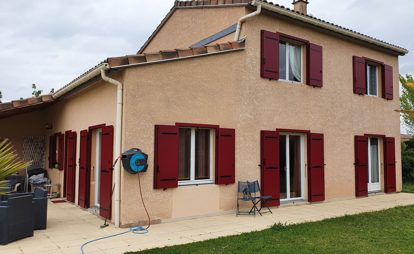 Une maison beige à deux étages avec un toit en tuiles de terre cuite, de grands volets rouges aux fenêtres et une terrasse donnant sur une pelouse.