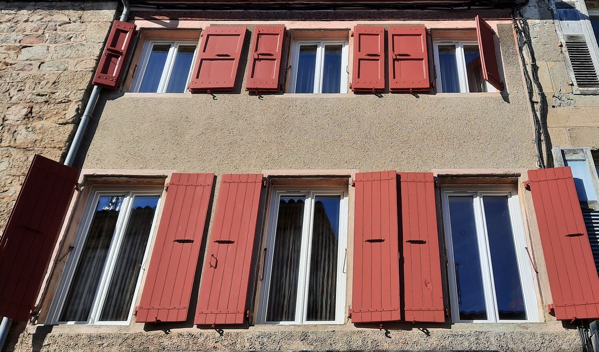 La façade d'un bâtiment de deux étages avec des murs en stuc beige et six fenêtres avec des volets en bois rouge rustiques ouverts.