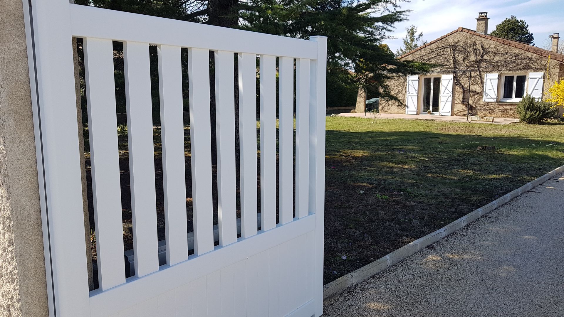 Au premier plan, un portail blanc à claire-voie se dresse, avec une maison en pierre et une pelouse à l'arrière-plan, sous un ciel bleu.