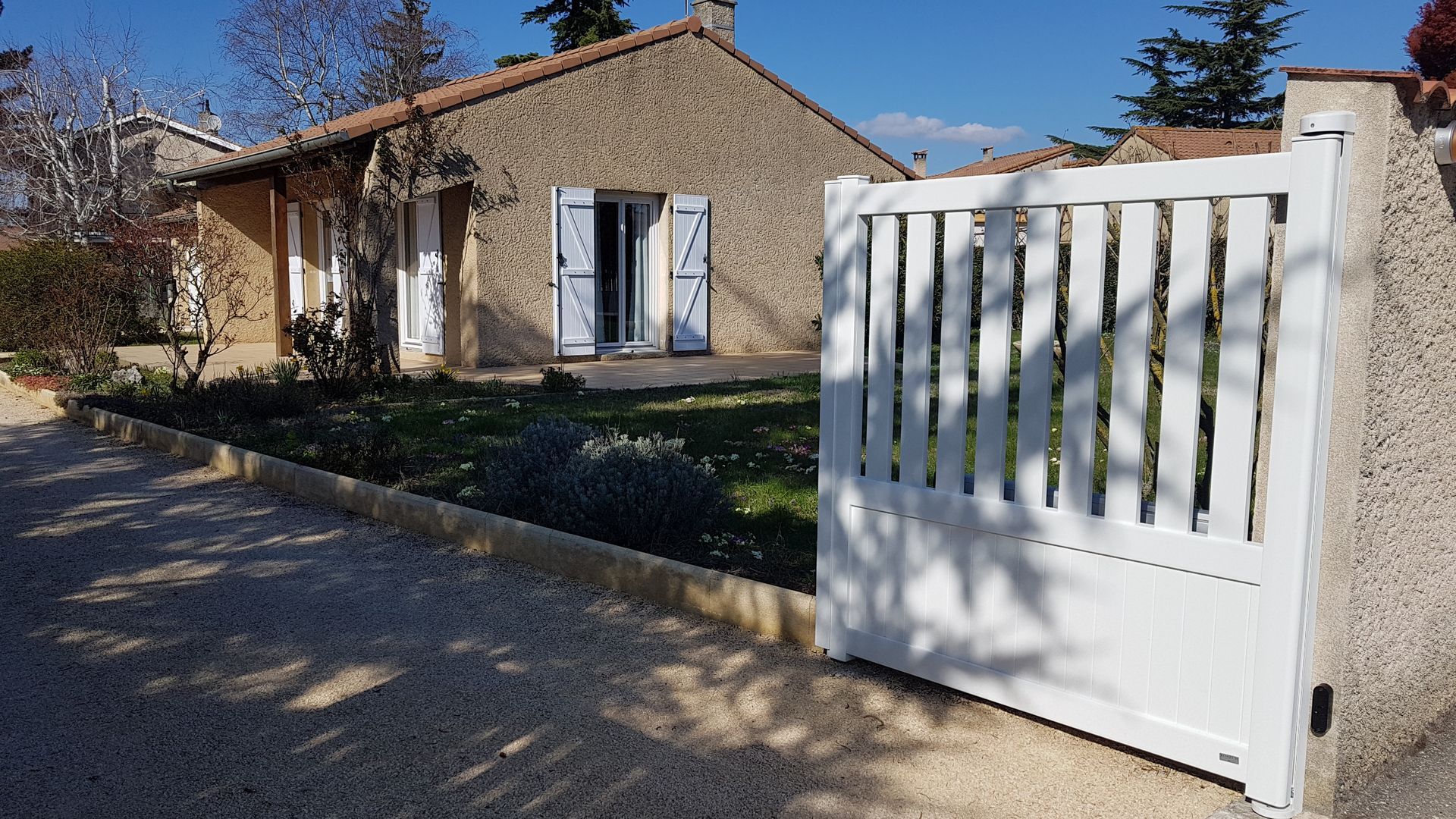 Une maison aux murs de pierre, avec des volets bleus et un portail blanc, dans une cour lumineuse et ensoleillée.