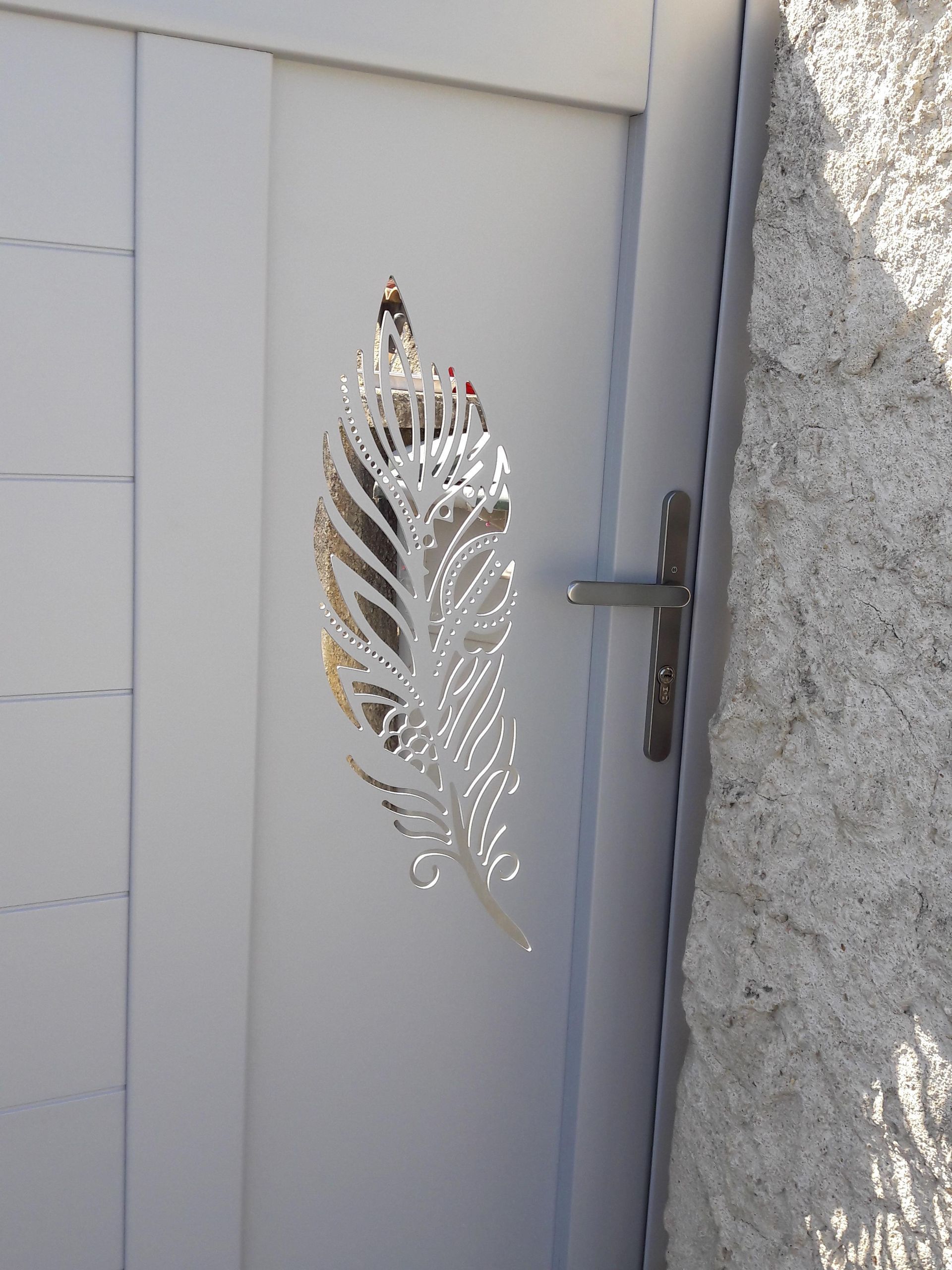 Une décoration décorative en métal blanc en forme de plume est accrochée à un portail gris, à côté d'un mur de pierre.