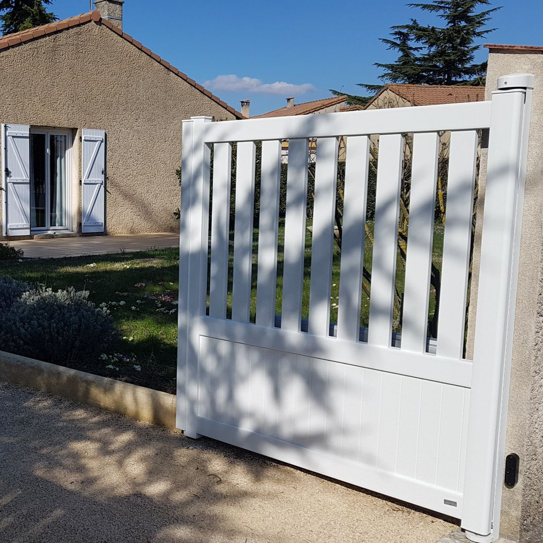 Un portail blanc à lattes verticales se dresse dans une allée de gravier devant une maison en pierre aux volets blancs.