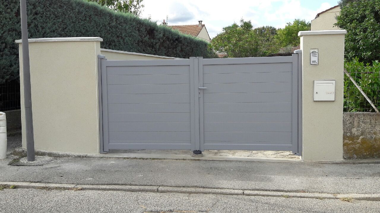 Un portail gris à double battant, encadré par des piliers en béton clair, avec une boîte aux lettres et un interphone sur le pilier de droite.