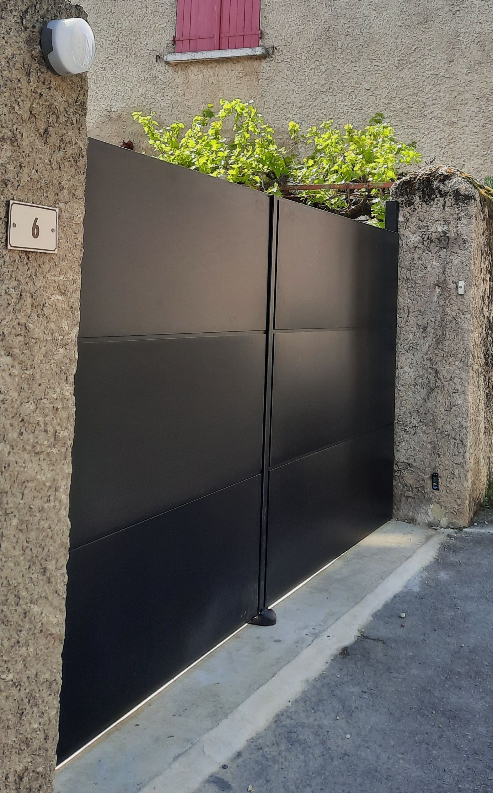 Un portail double moderne en métal noir massif, encadré par deux piliers en pierre texturée, vu depuis la rue.