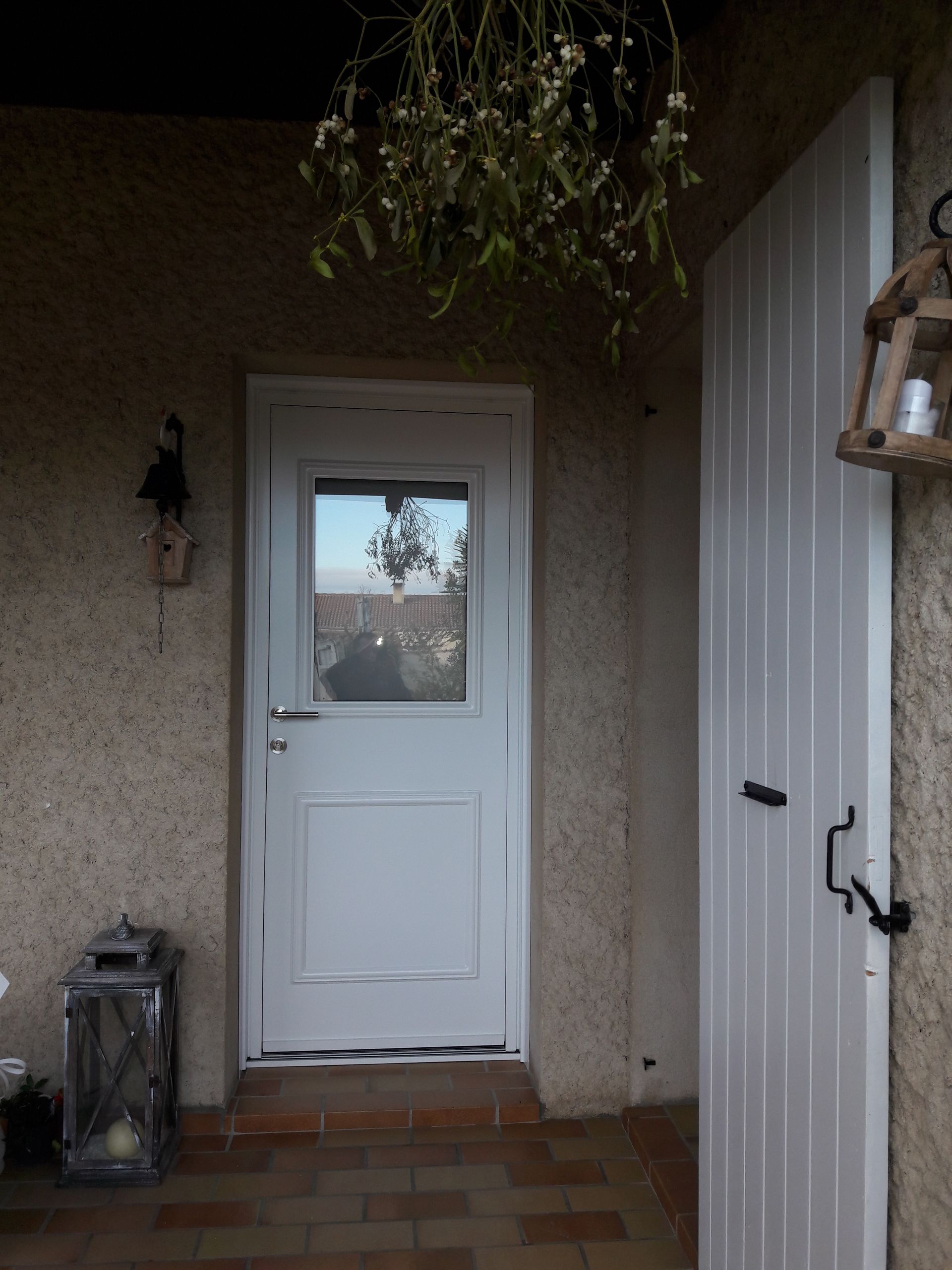 Une porte blanche et un panneau latéral sous un porche couvert orné de plantes suspendues, d'une applique murale et d'une lanterne sur pied.