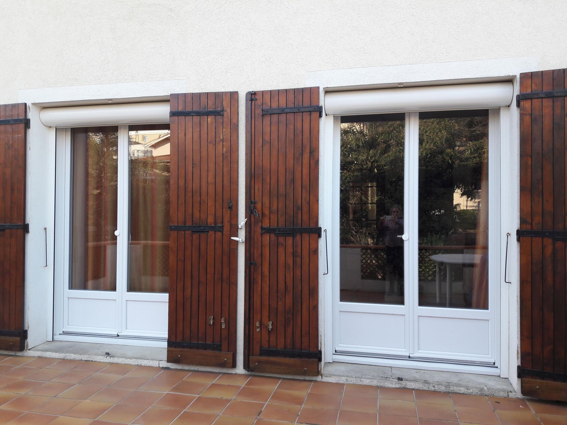 Deux portes-fenêtres à encadrement blanc et volets en bois foncé donnent sur une terrasse carrelée.