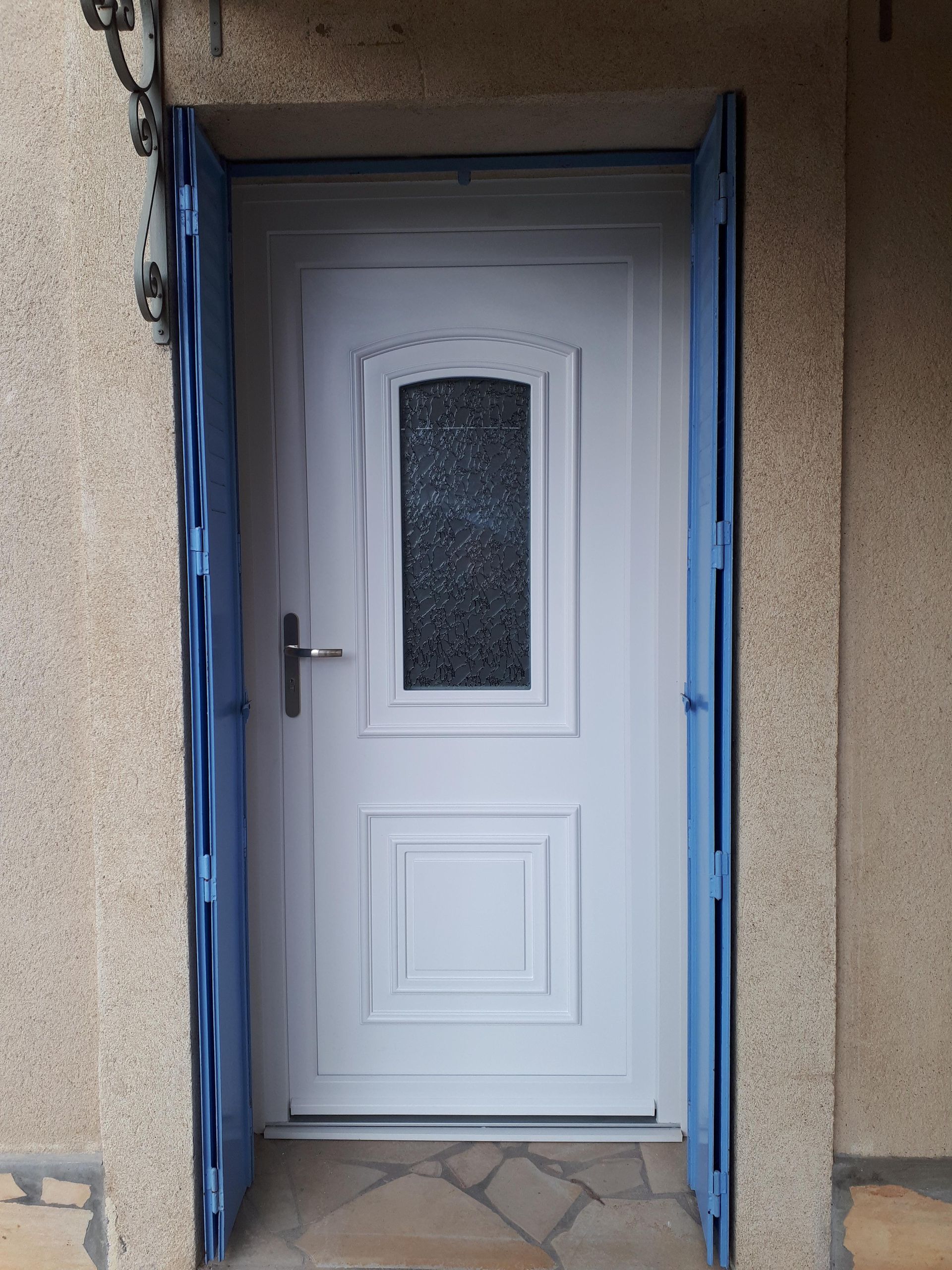Une porte blanche avec une petite fenêtre en verre texturé, encadrée par des grilles de sécurité bleues sur une façade en pierre texturée.